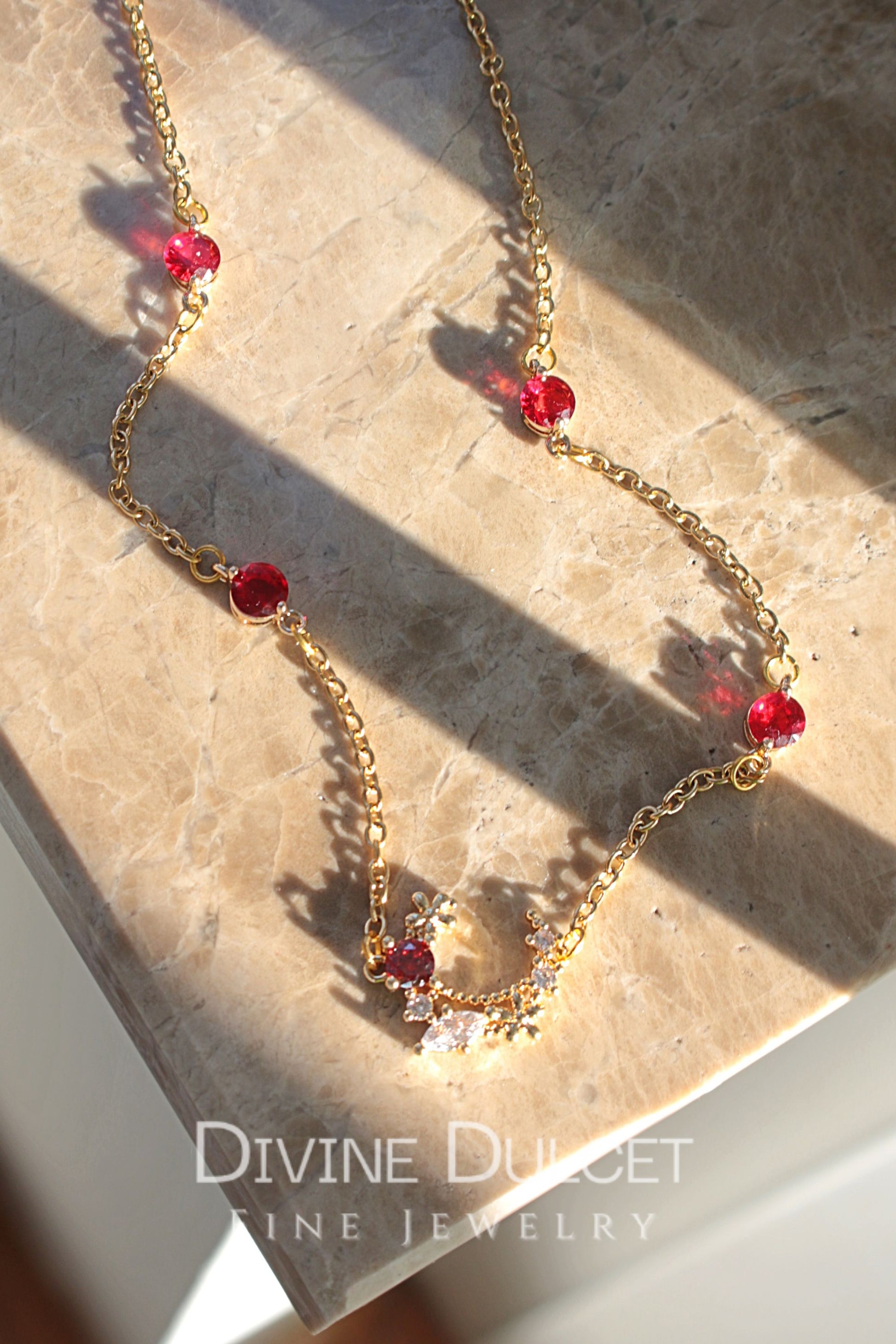 Gold necklace with red beads on a stone surface, featuring 'Divine Dulcet Fine Jewelry' branding.