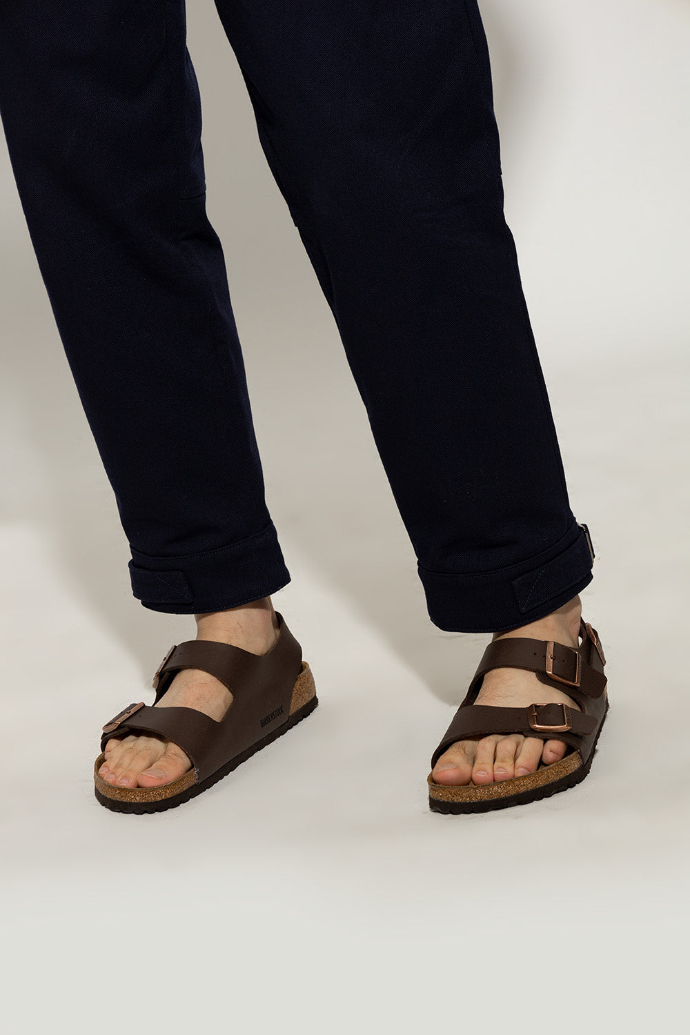 Person wearing dark brown sandals and black pants on a white background