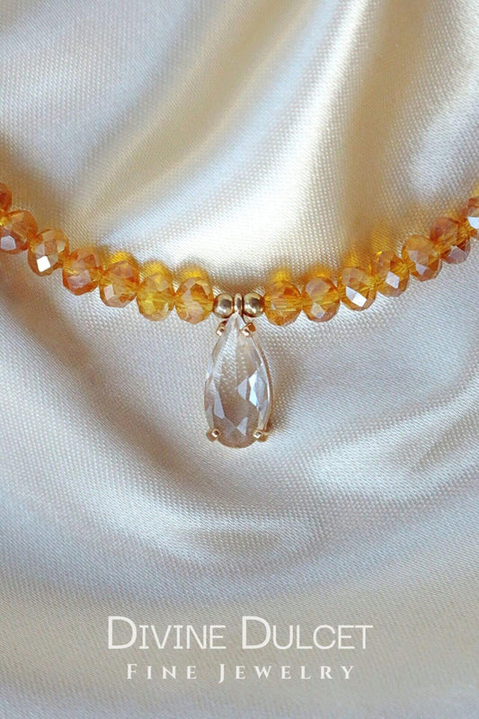 Necklace with amber beads and a clear teardrop pendant on a satin background, branded 'Divine Dulcet'.