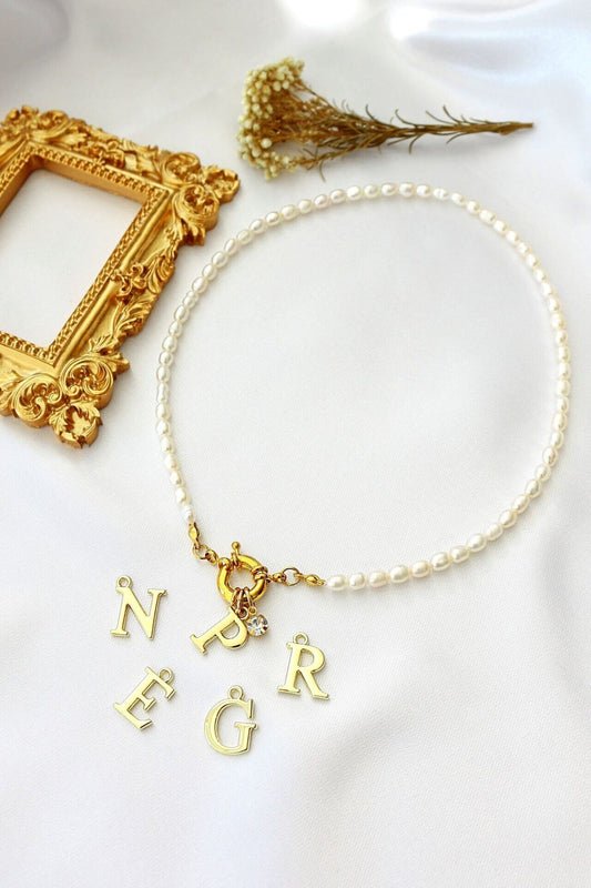Pearl necklace with gold letter charms on a white surface with a gold frame and dried flowers.
