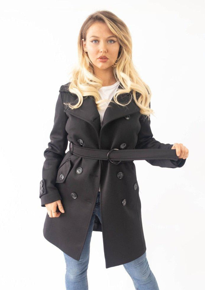 Woman wearing a black trench coat with a belt, standing against a white background.