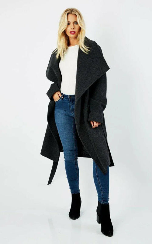 Woman wearing a long black coat, white top, blue jeans, and black boots on a white background