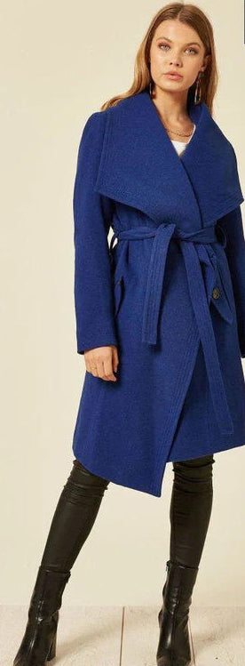 Woman wearing a blue coat with a belt, standing against a plain background.