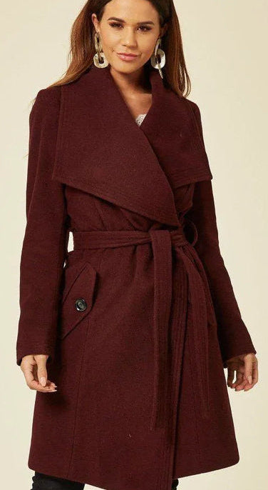 Woman wearing a burgundy coat with a belt, standing against a beige background. 