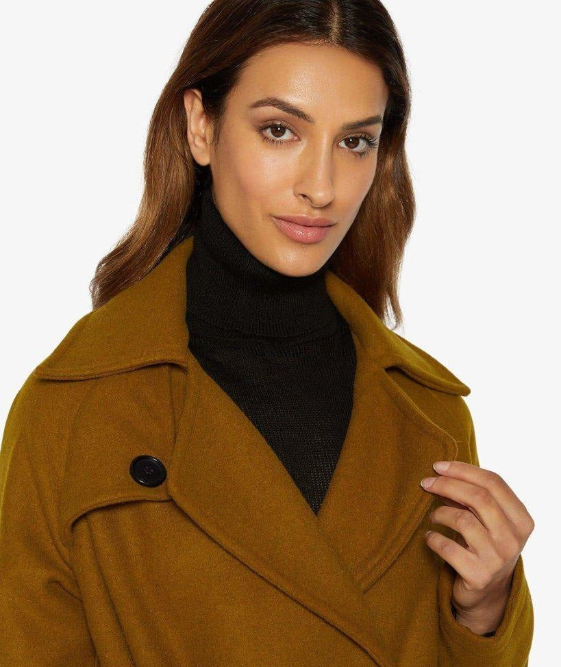 Woman wearing a mustard yellow coat over a black turtleneck on a white background