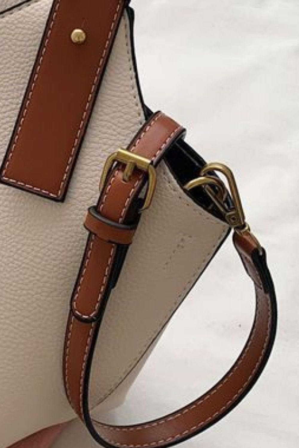 Close-up of a beige handbag with brown leather strap and gold buckle on a white background