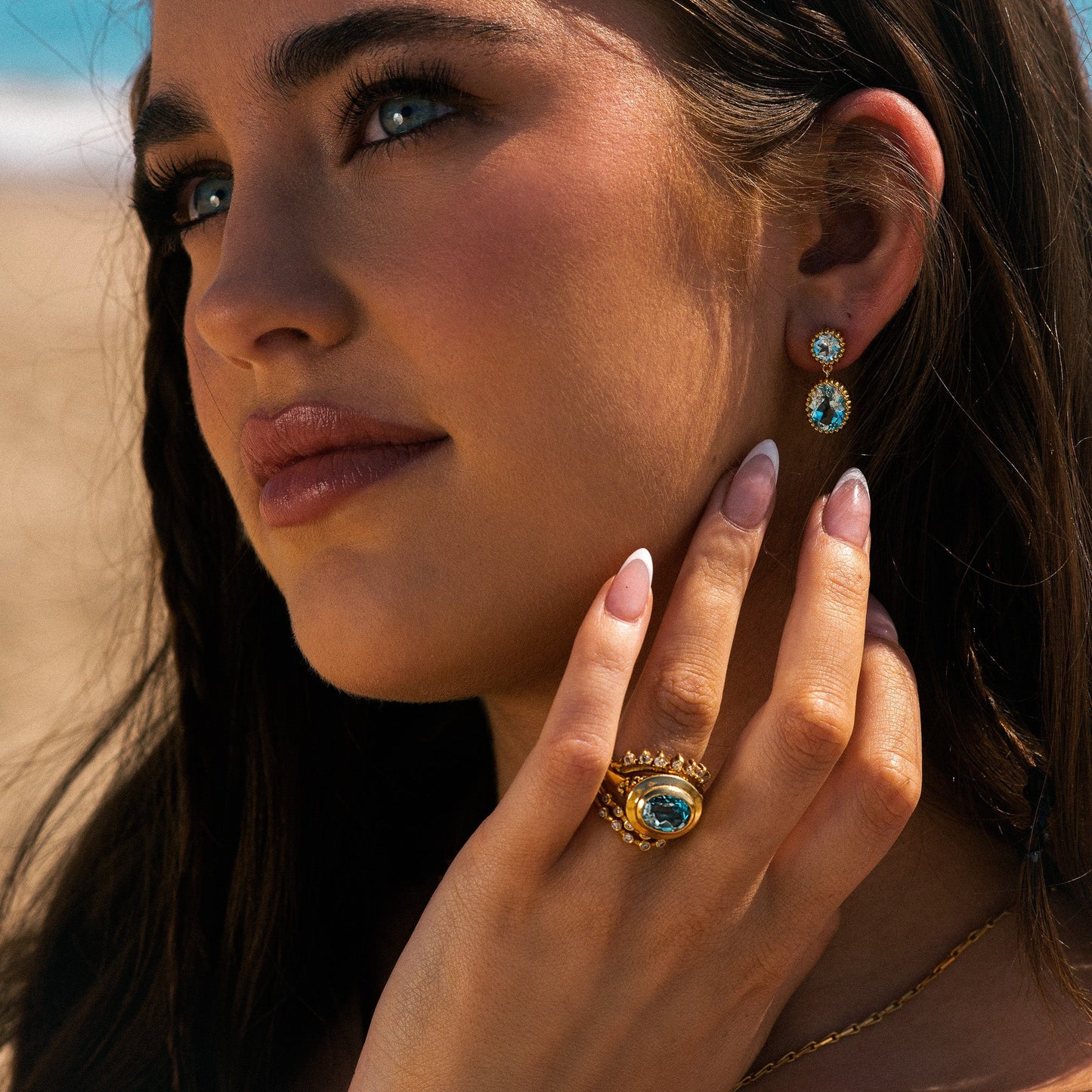 Close-up of a woman wearing gold earrings and ring with blue gemstones against a blurred background