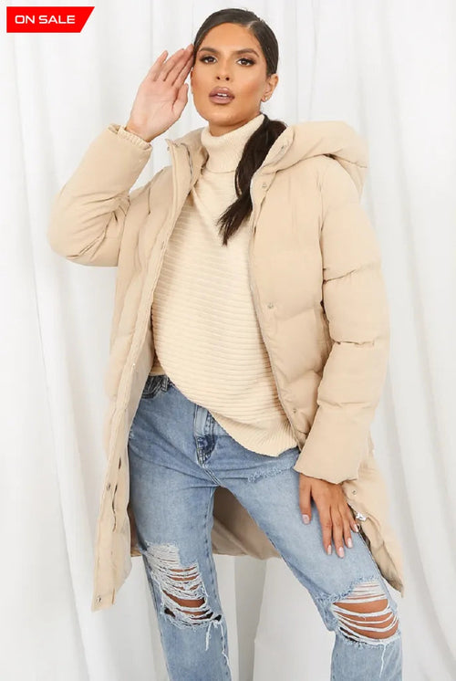 Woman wearing a beige puffer jacket, beige sweater, and ripped jeans on a white background.