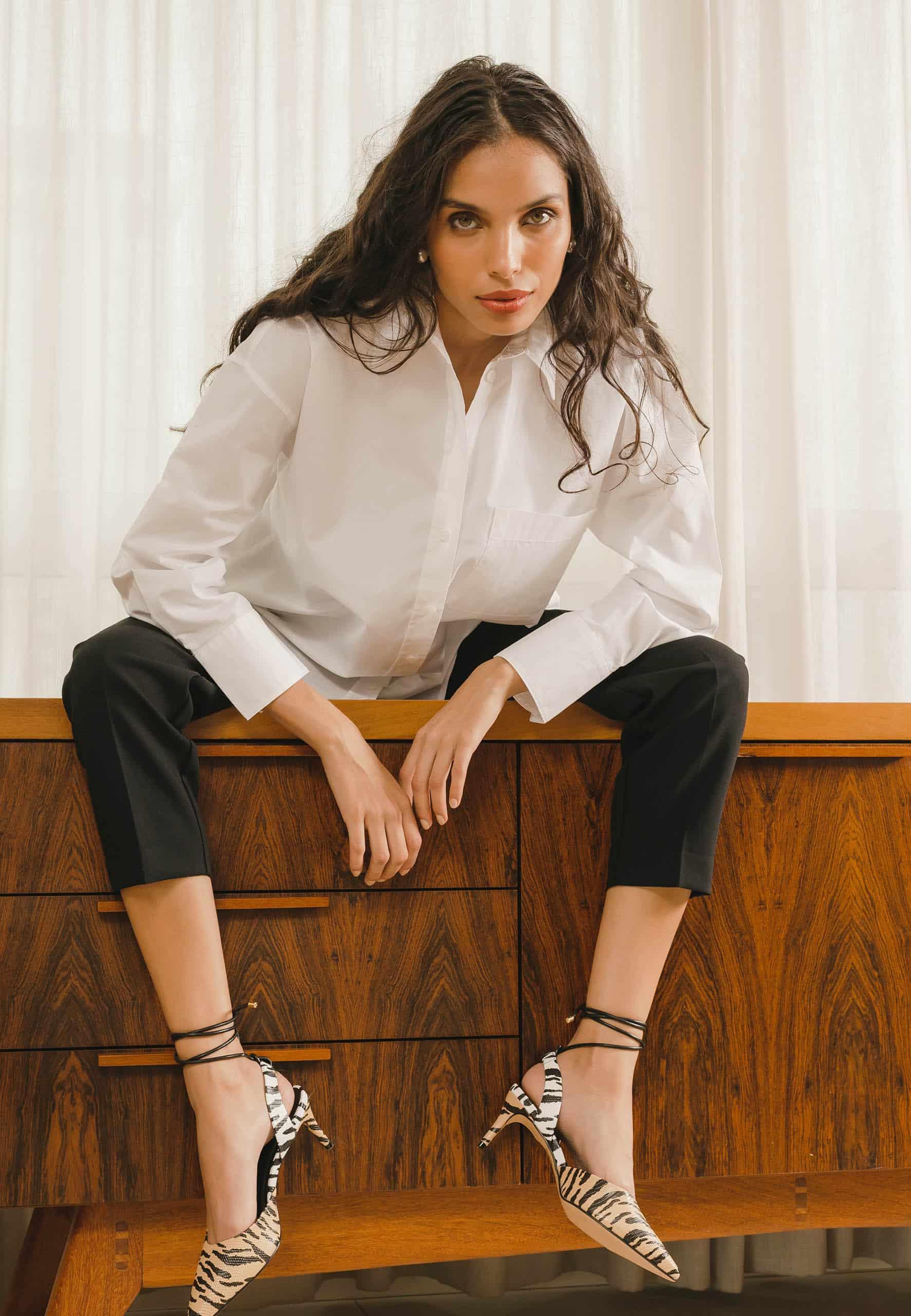 Woman sitting on a wooden dresser wearing a white shirt and black pants.