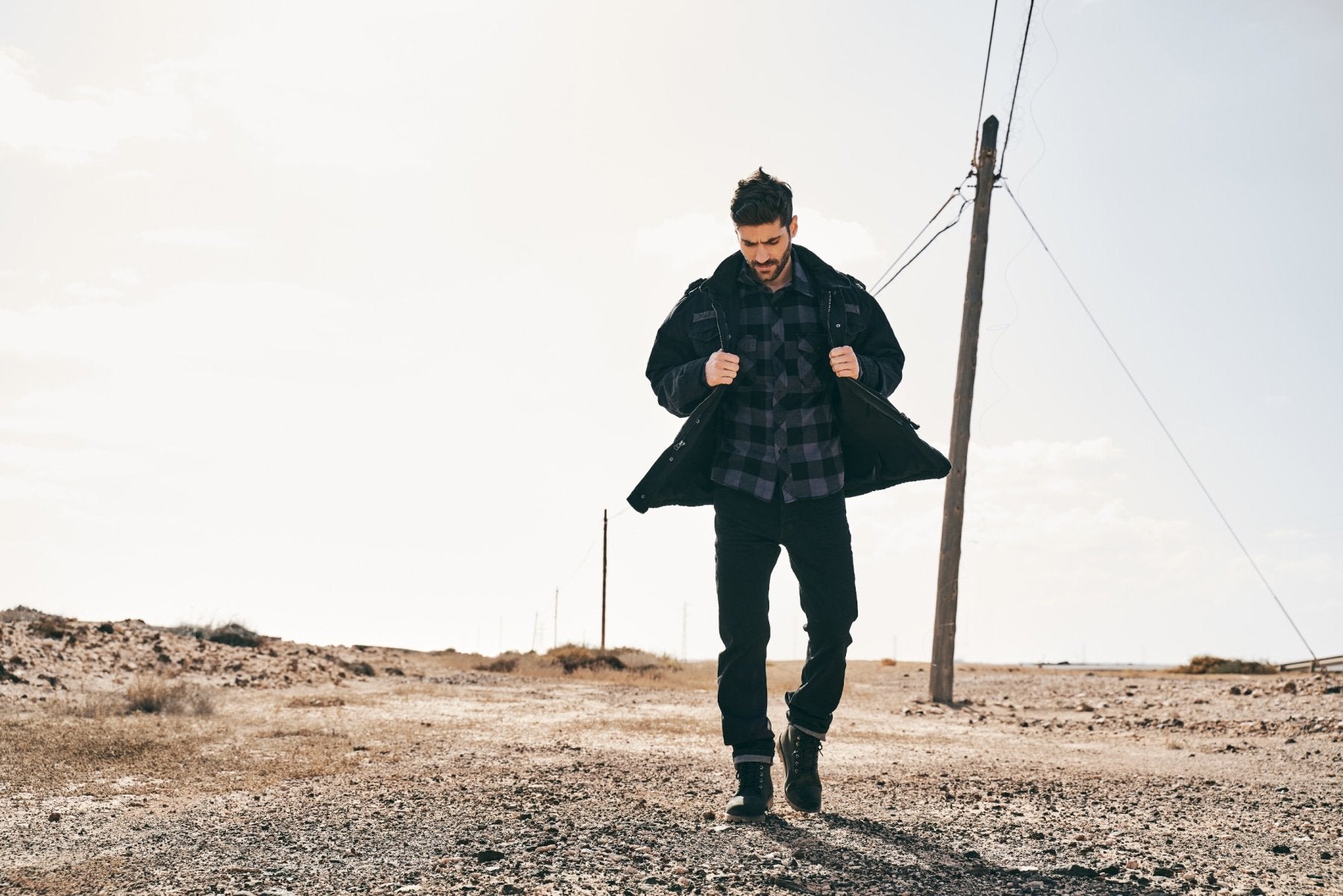 Man in a coat and plaid shirt standing in a desert-like landscape with power lines in the background.