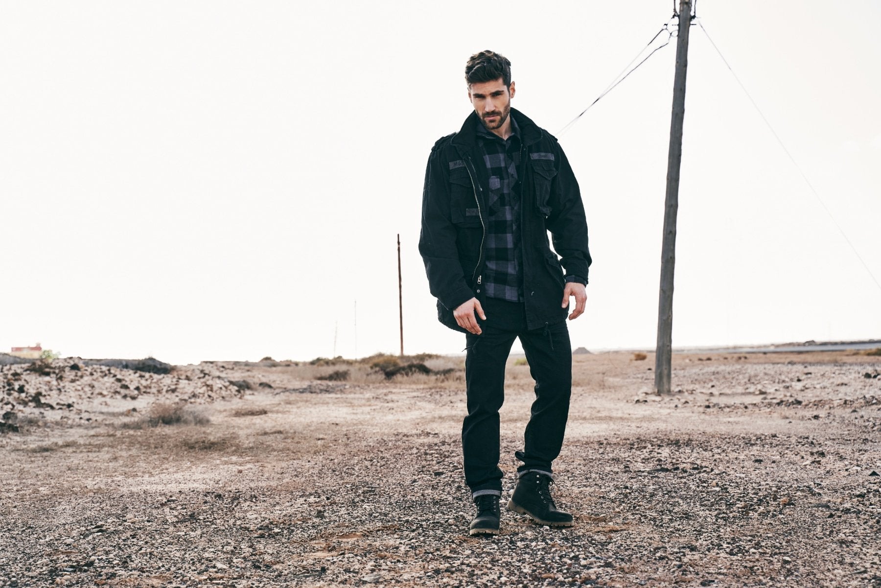 Man wearing a black coat and plaid shirt standing in a barren landscape.