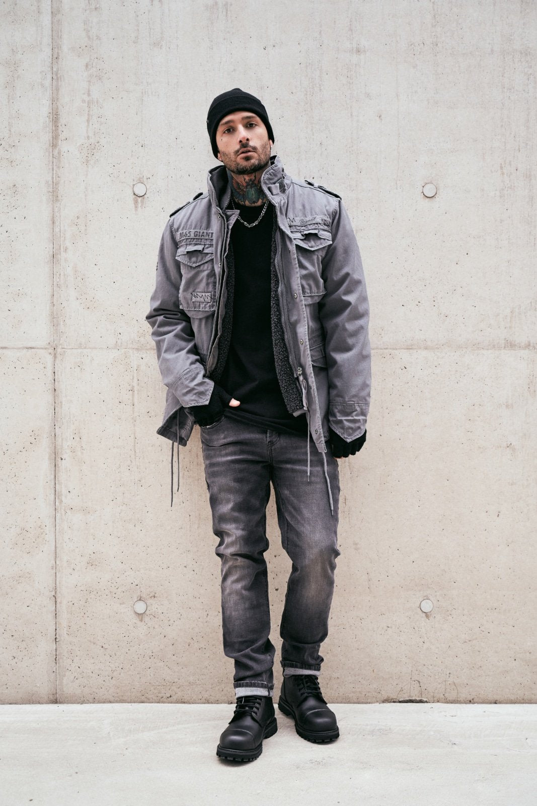 Person wearing a gray jacket, black beanie, and black shoes against a concrete wall.