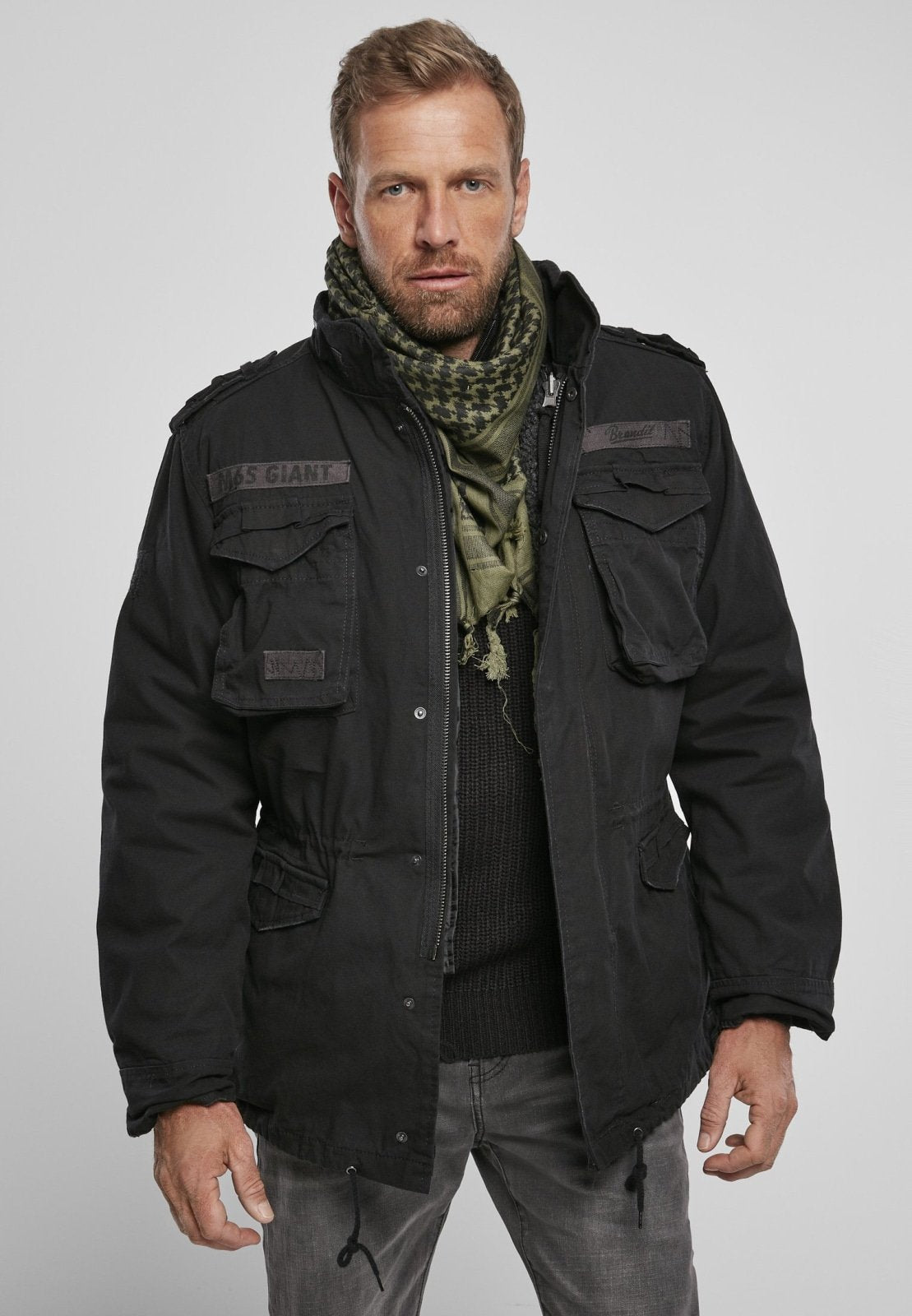 Man wearing a black jacket with multiple pockets and a patterned scarf on a gray background