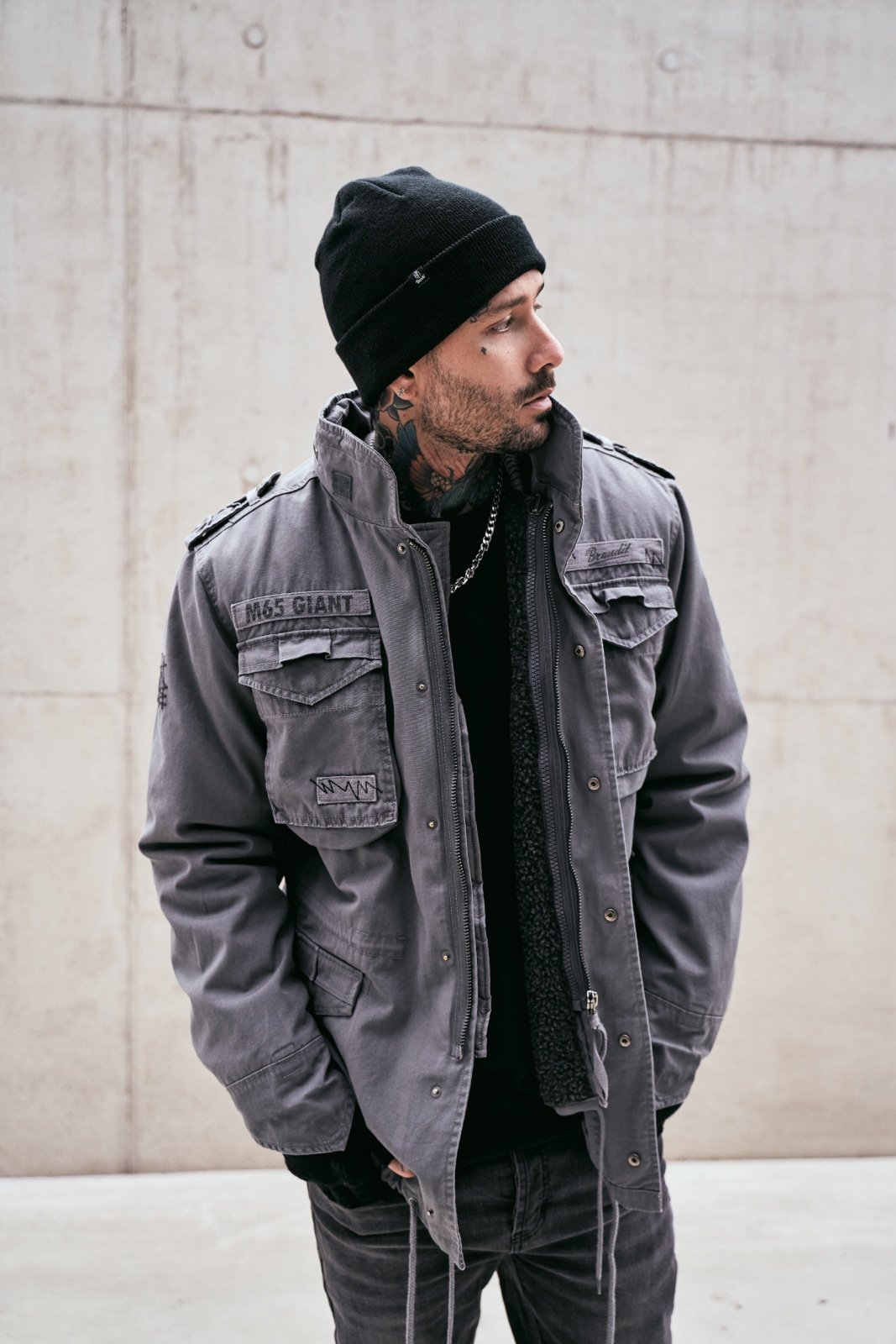 Man wearing a gray jacket and black beanie standing against a concrete wall.