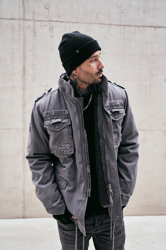 Man wearing a gray jacket and black beanie standing against a concrete wall.