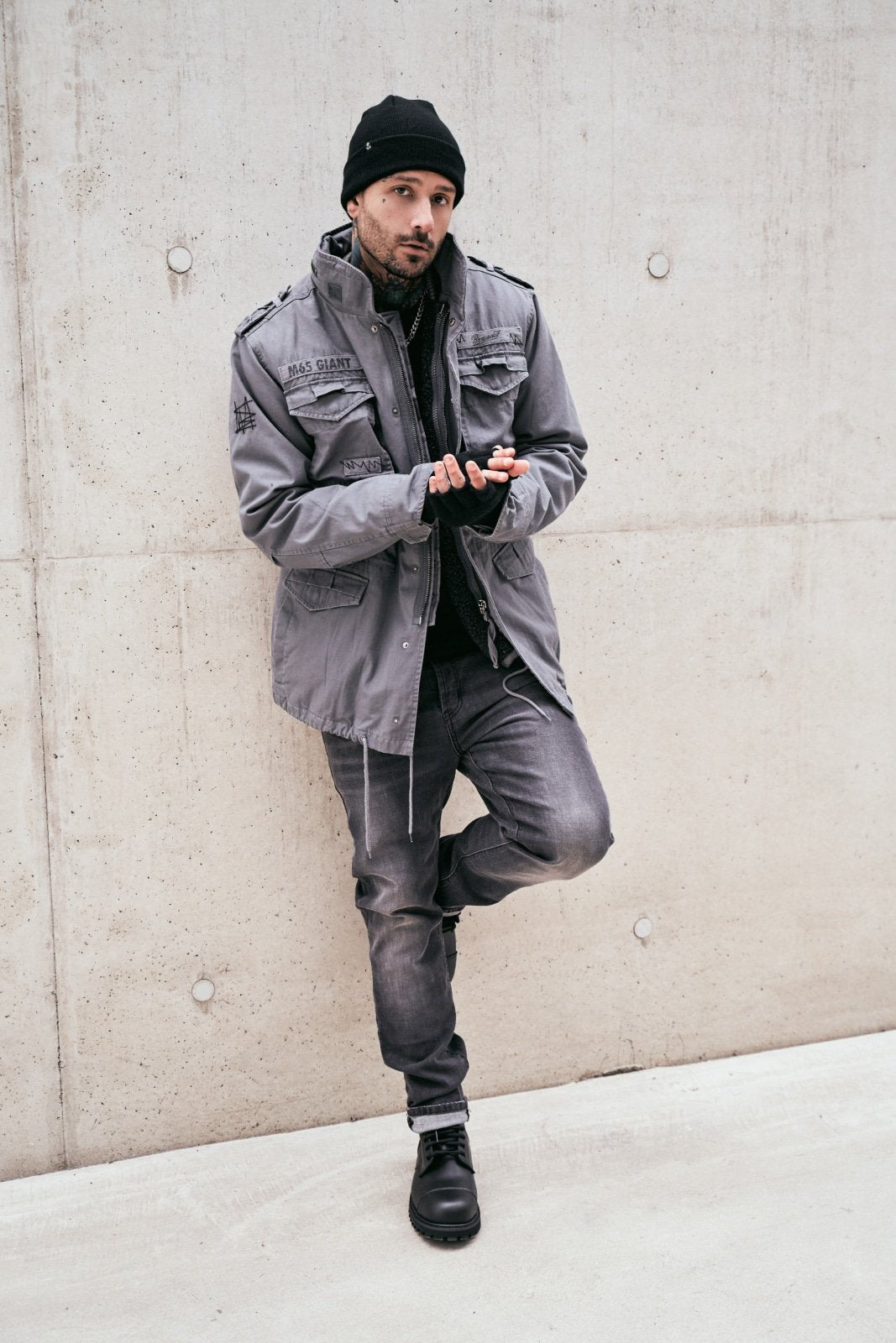 Man wearing a gray jacket and pants leaning against a concrete wall.