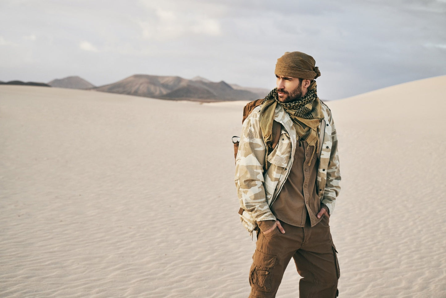 Man standing on a sand dune wearing a camouflage jacket and brown pants with mountains in the background.