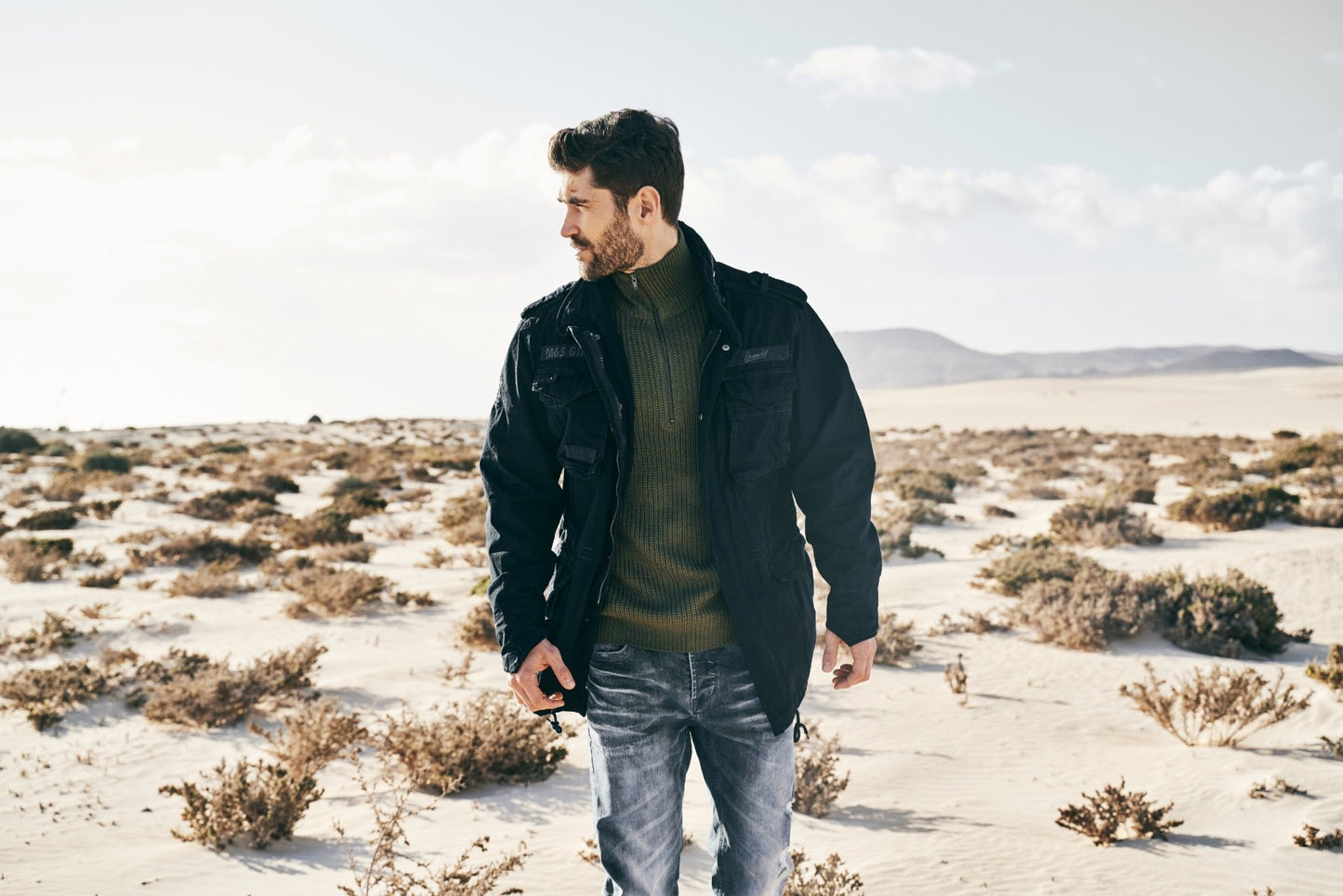 Man standing in a desert landscape wearing a dark jacket and jeans.