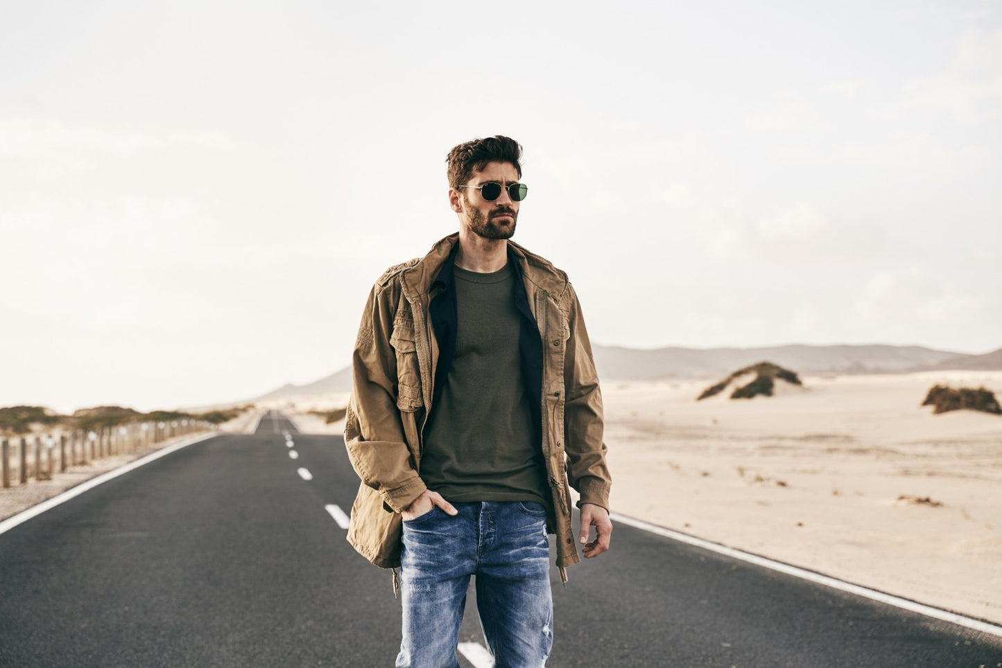 Man standing on a desert road wearing sunglasses and a jacket.