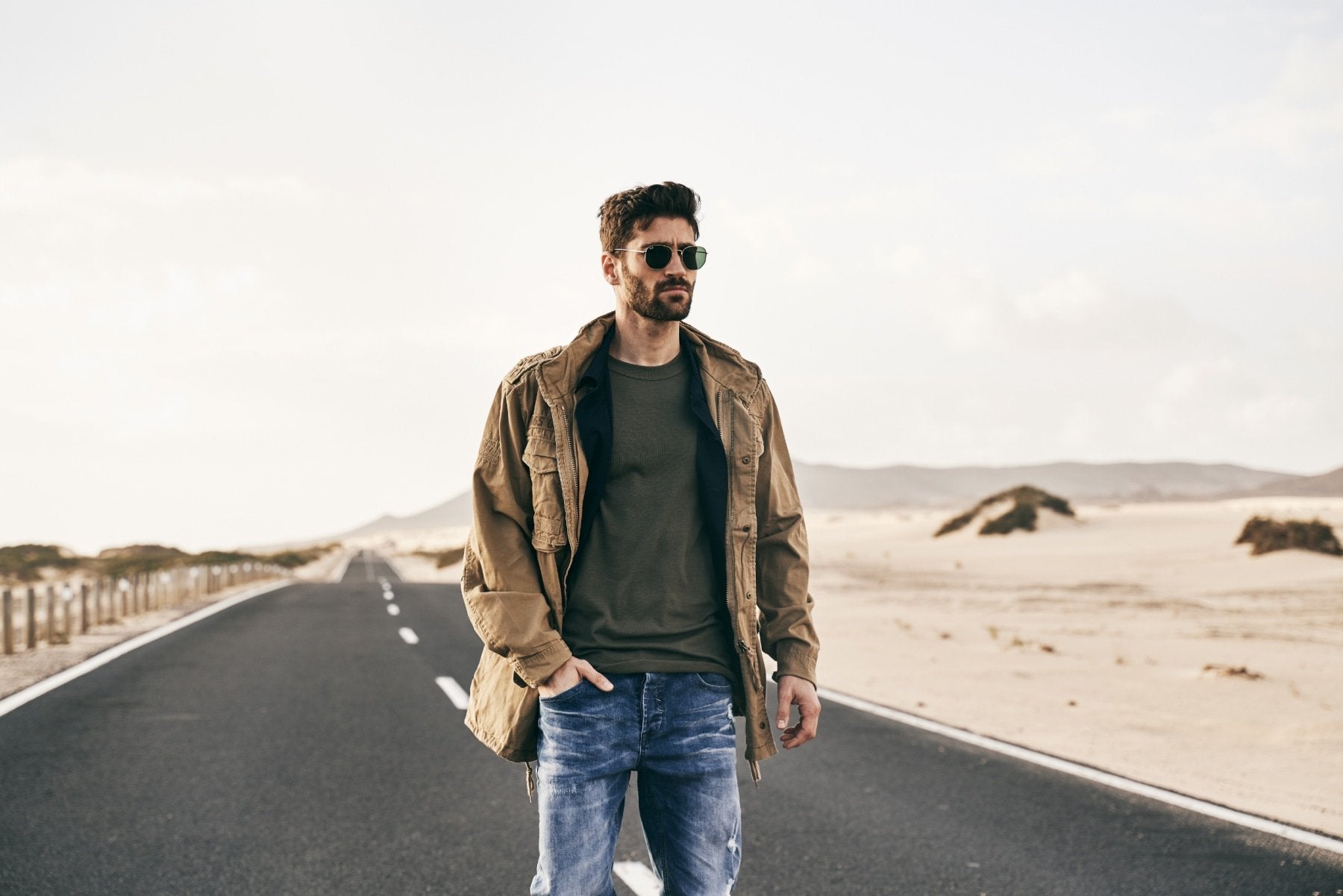 Man standing on a desert road wearing sunglasses and a jacket.