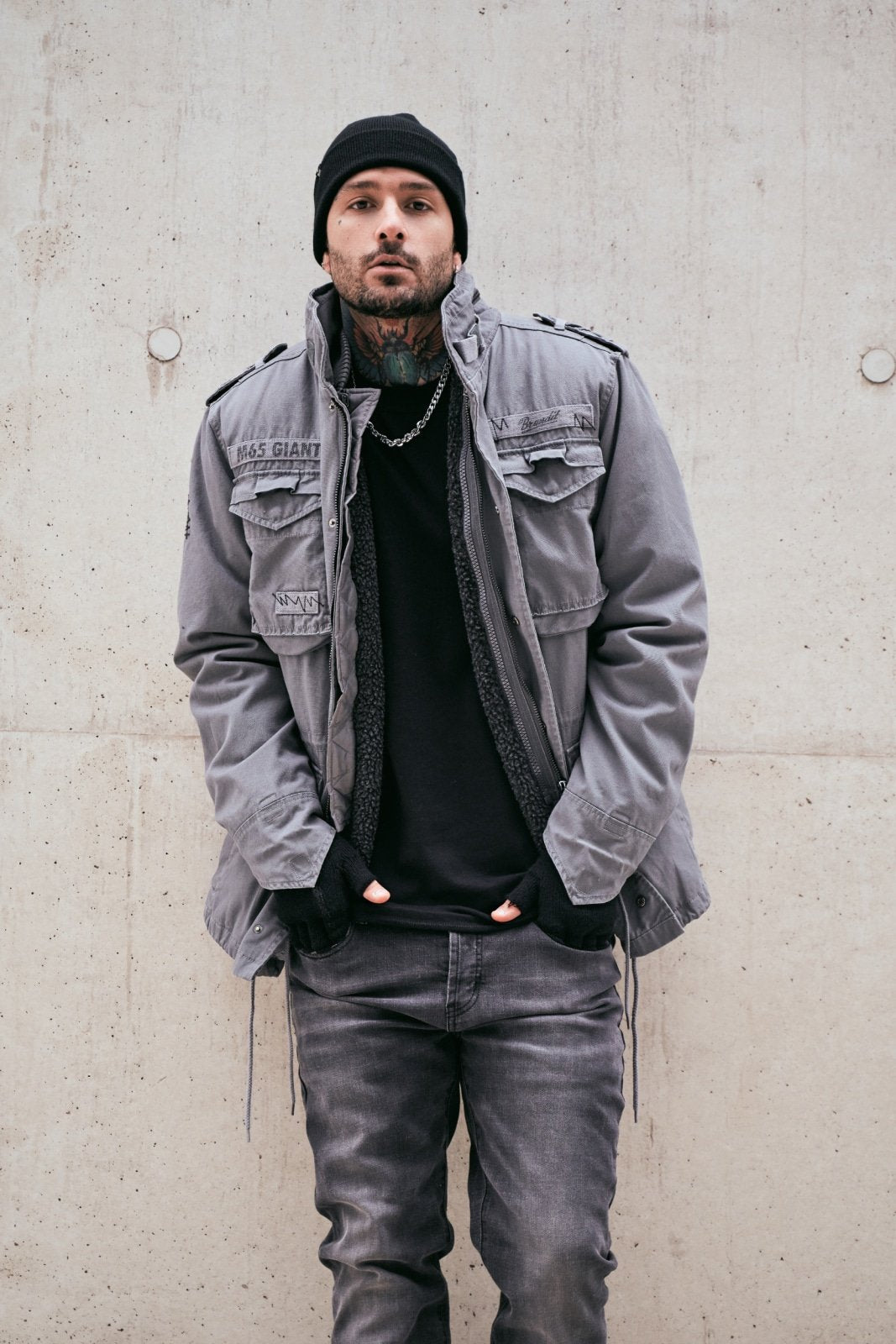 Man wearing a gray jacket and black beanie standing against a concrete wall.