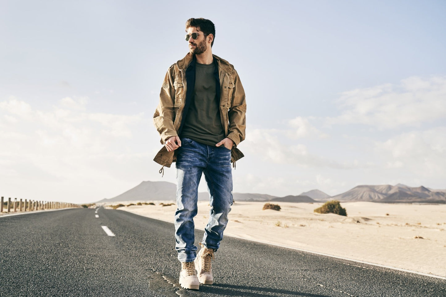 Man standing on a desert road wearing sunglasses, a jacket, and jeans.