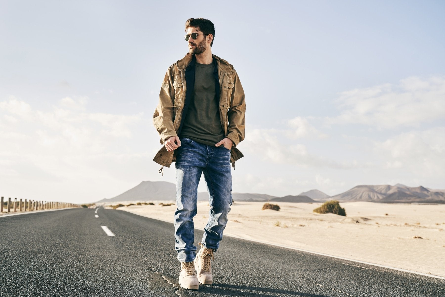 Man standing on a desert road wearing sunglasses, a jacket, and jeans.