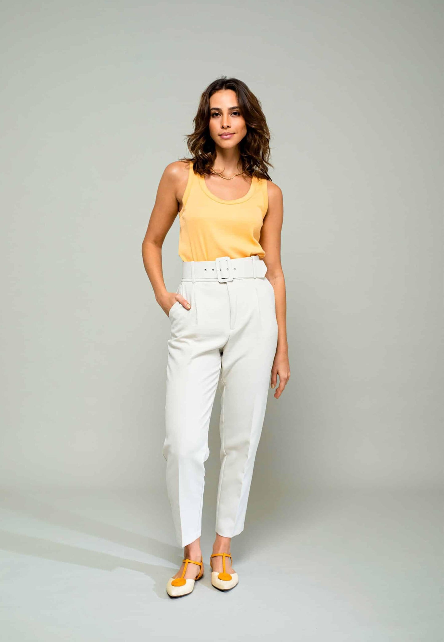Woman wearing a yellow sleeveless top and white pants against a gray background