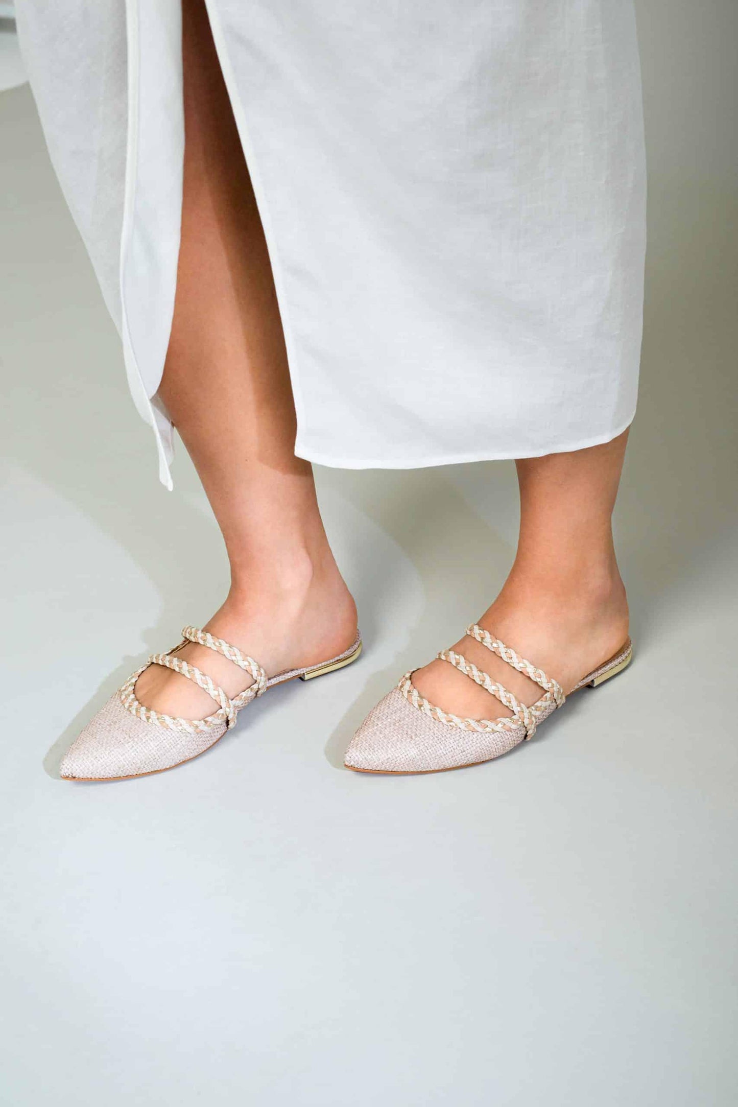 White sandals with thin straps worn by a person in a white skirt on a light gray background