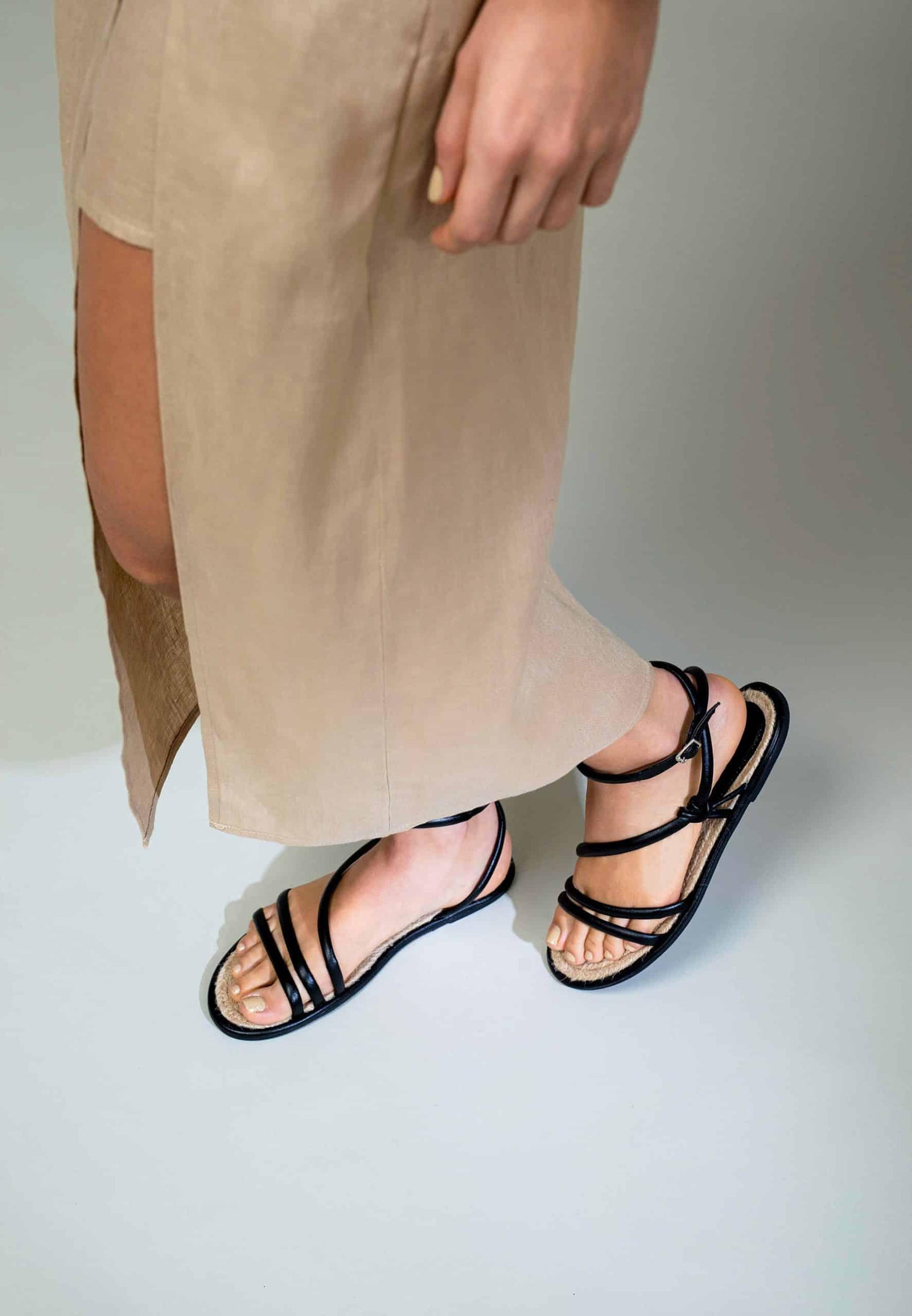 Person wearing black strappy sandals with a beige skirt on a light gray background