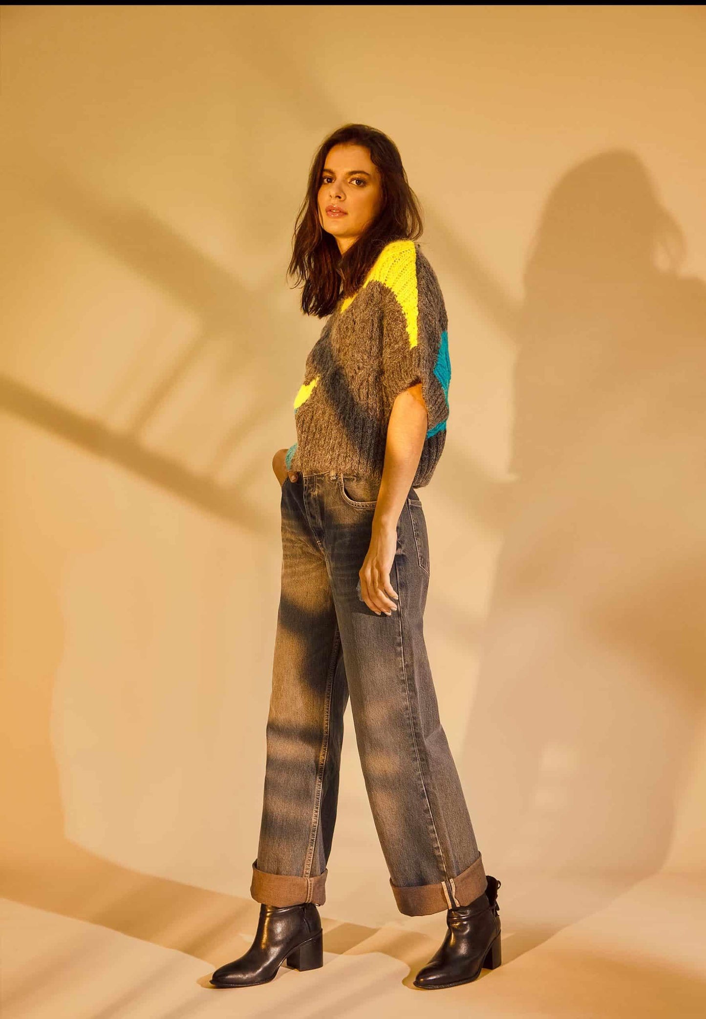 Woman wearing a patterned sweater and jeans standing against a beige wall.