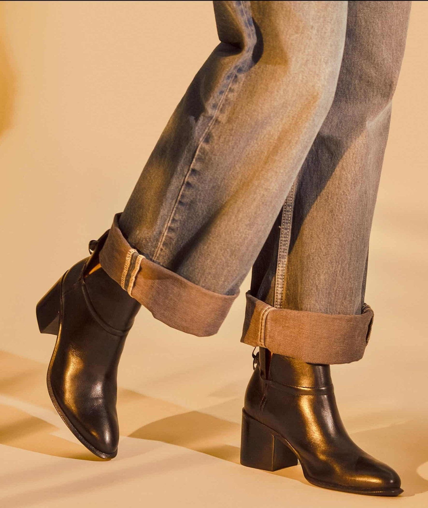 Black leather ankle boots worn with rolled-up jeans on a beige background