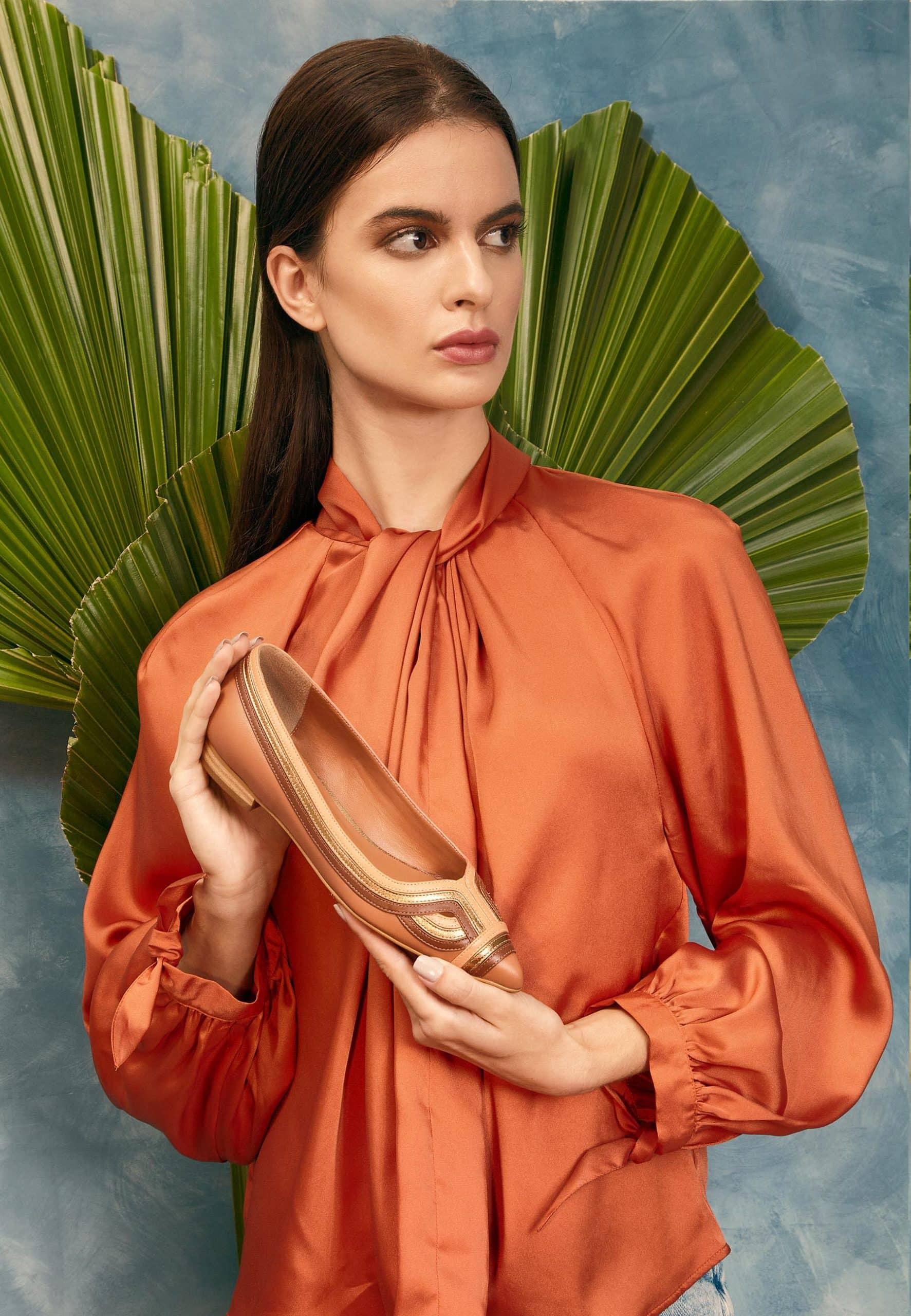 Woman in an orange blouse holding a shoe against a blue background with palm leaves