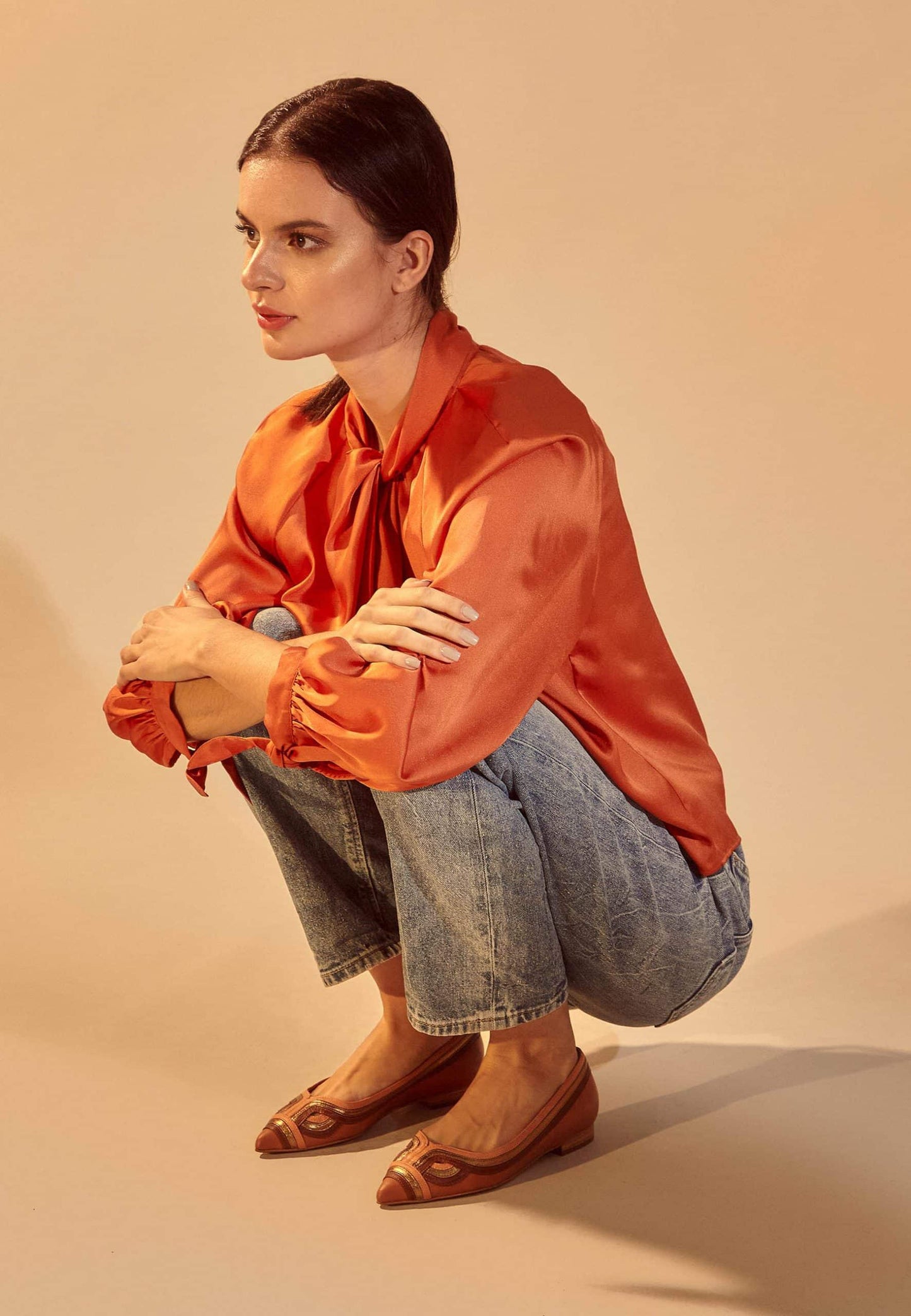 Woman wearing an orange satin blouse and denim pants on a beige background