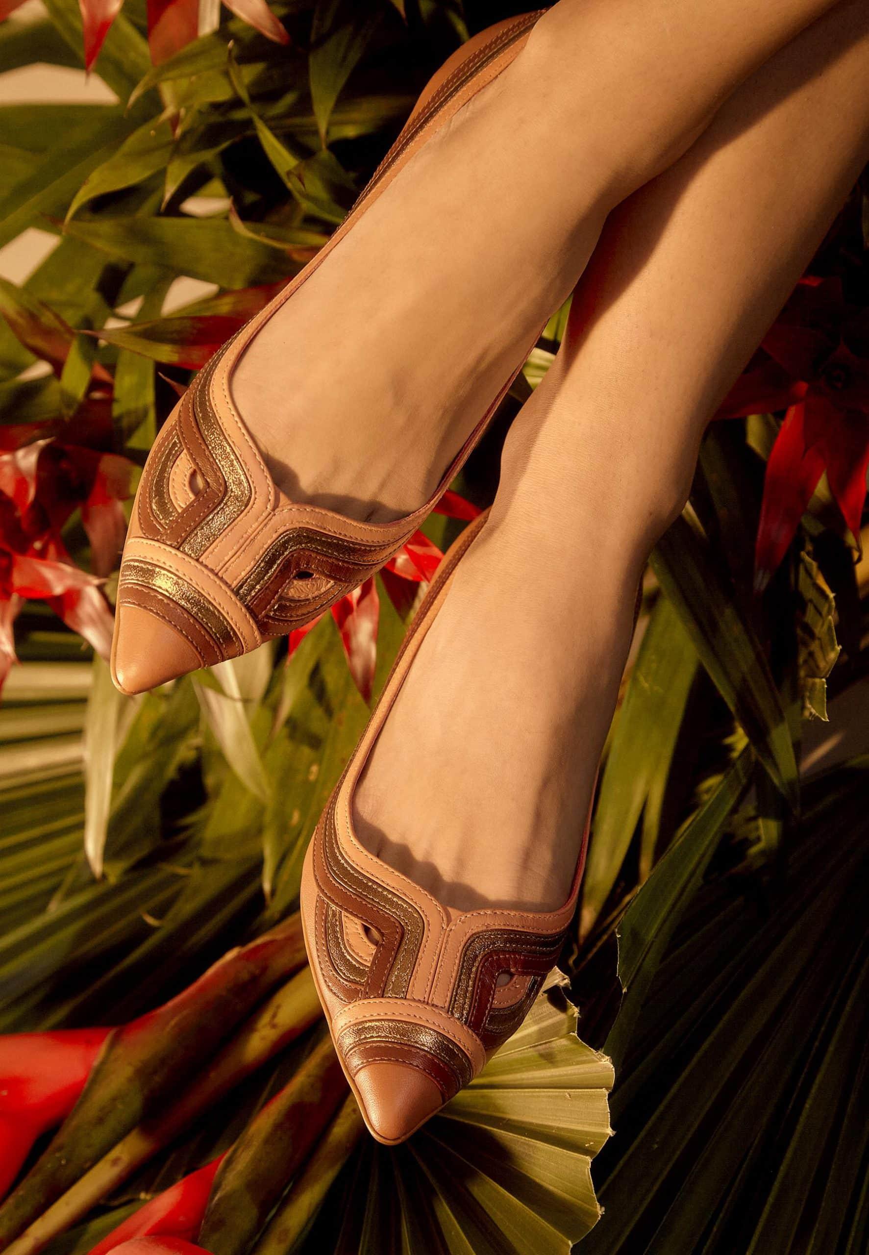 Brown pointed-toe shoes with decorative patterns worn by a person against a tropical plant background.