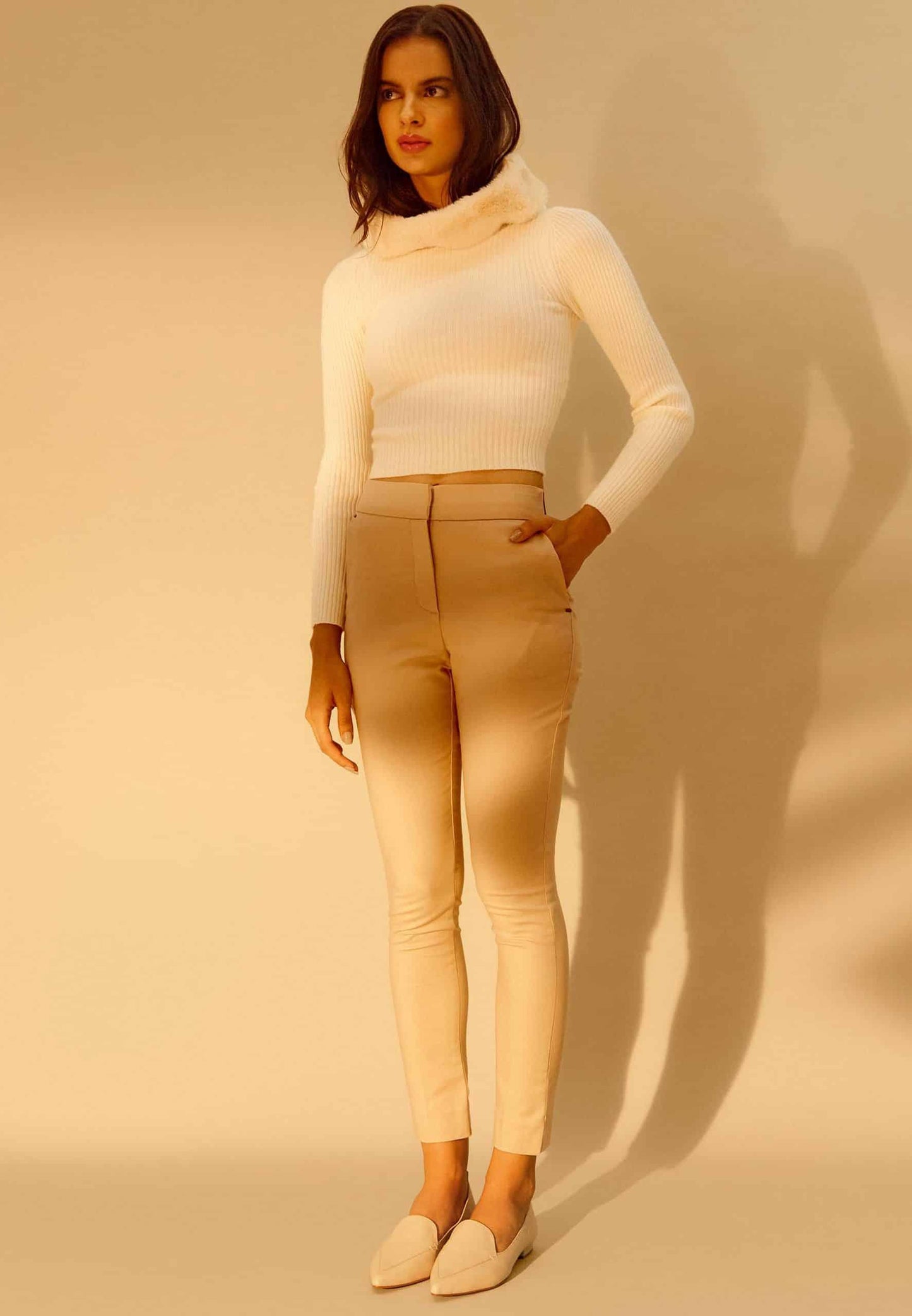 Woman wearing a white turtleneck and beige pants against a beige background