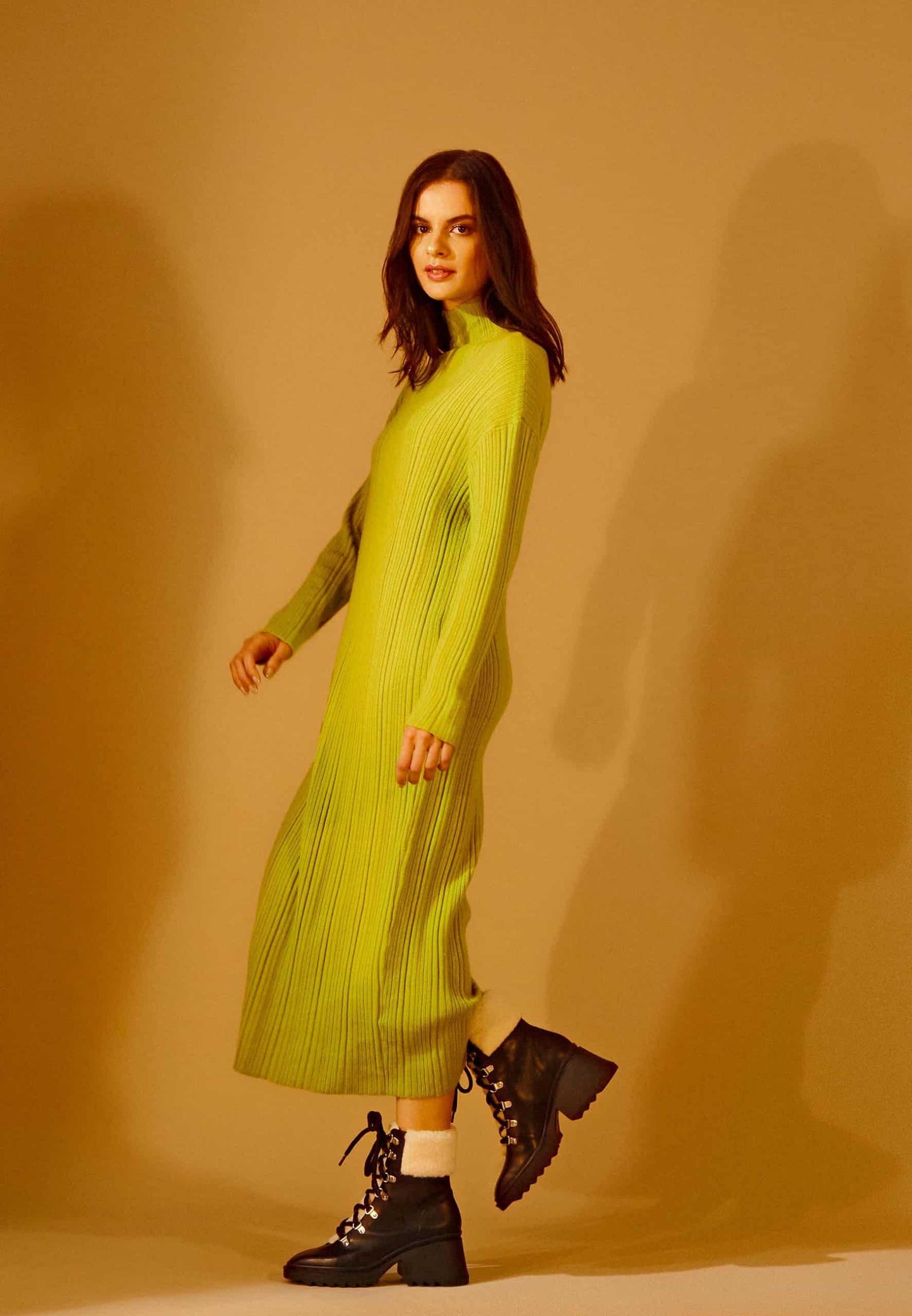 Woman wearing a green pleated dress and black boots against a beige background