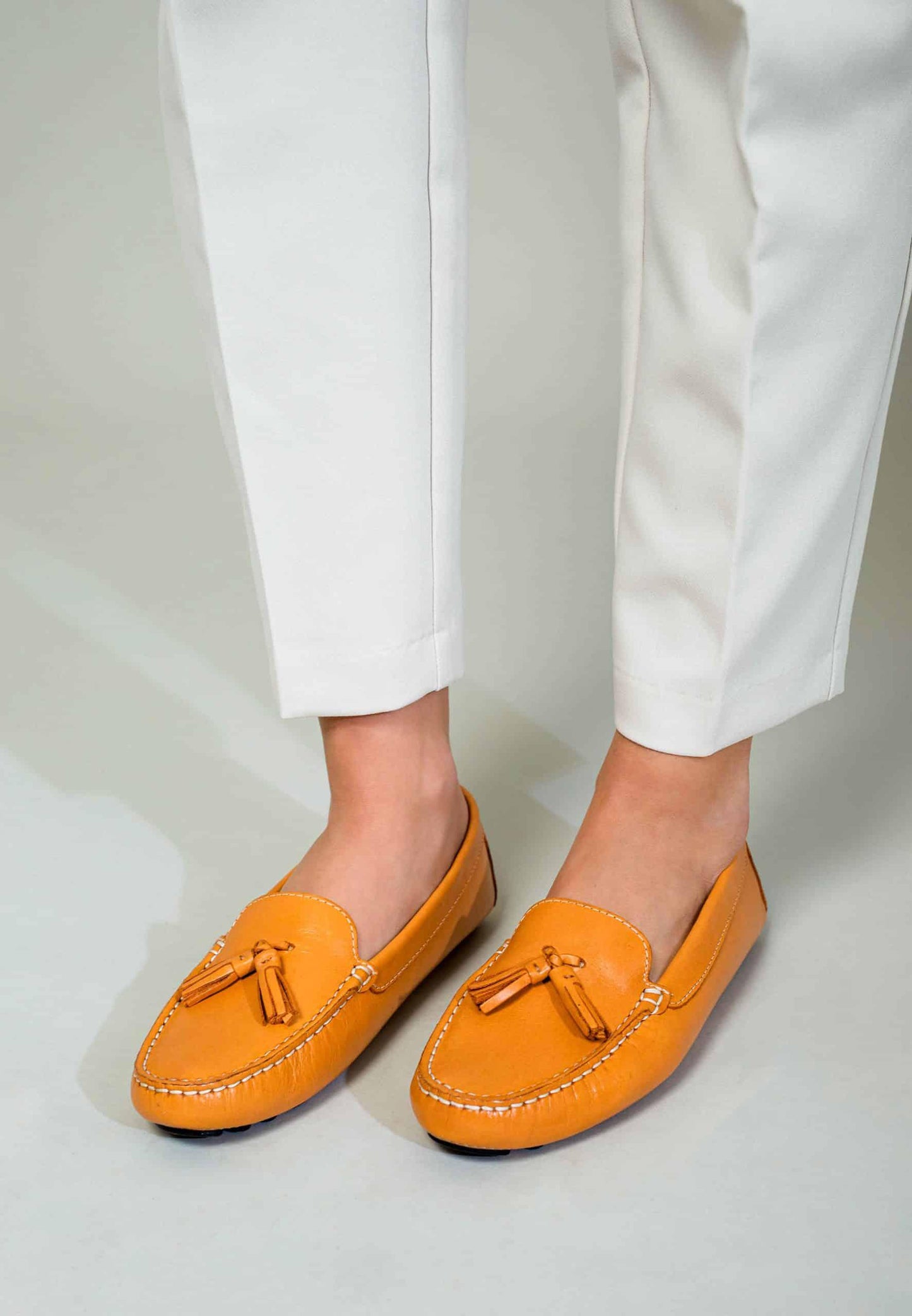 Orange loafers with tassels worn with white pants on a light gray background