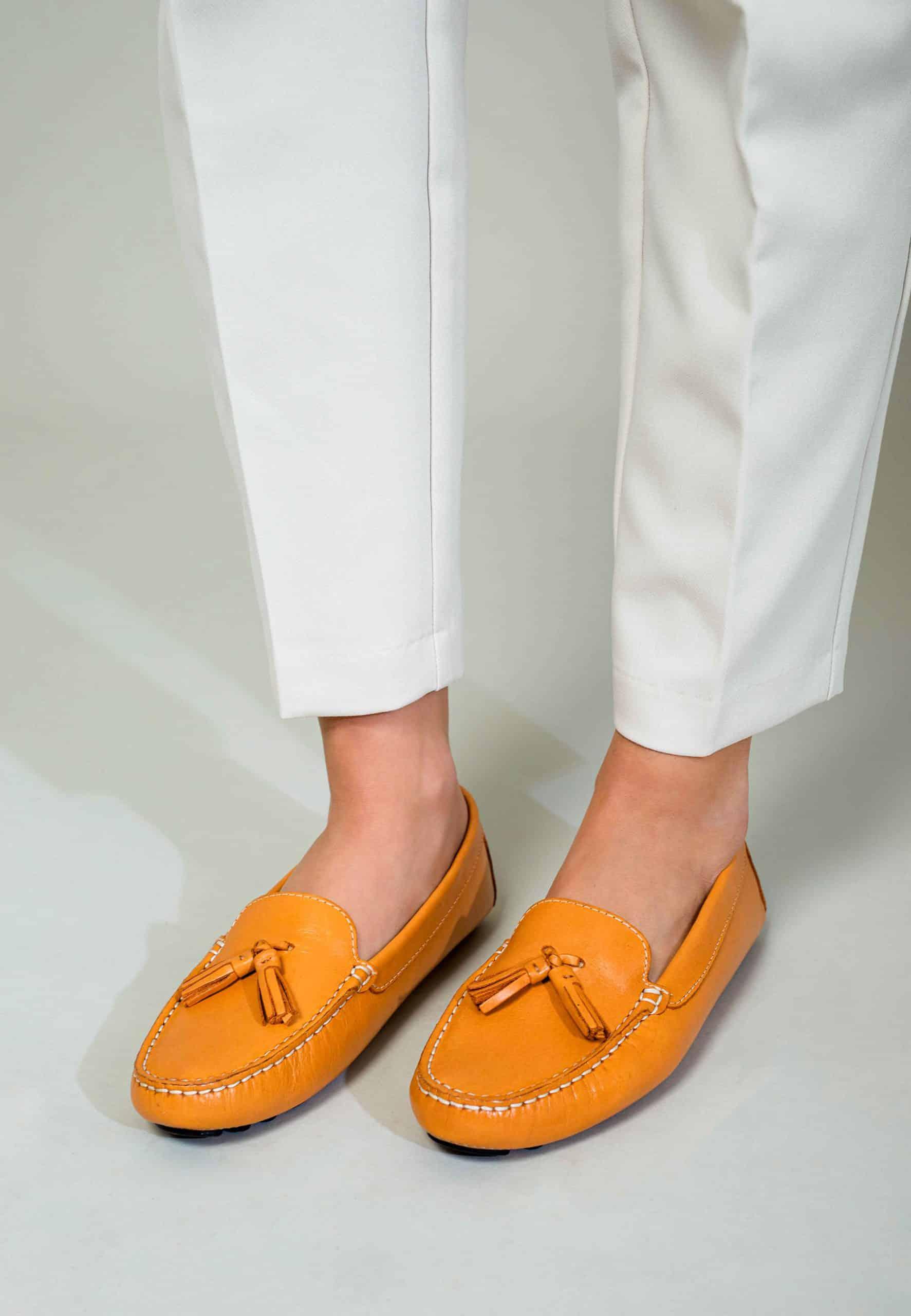 Orange loafers with tassels worn with white pants on a light gray background