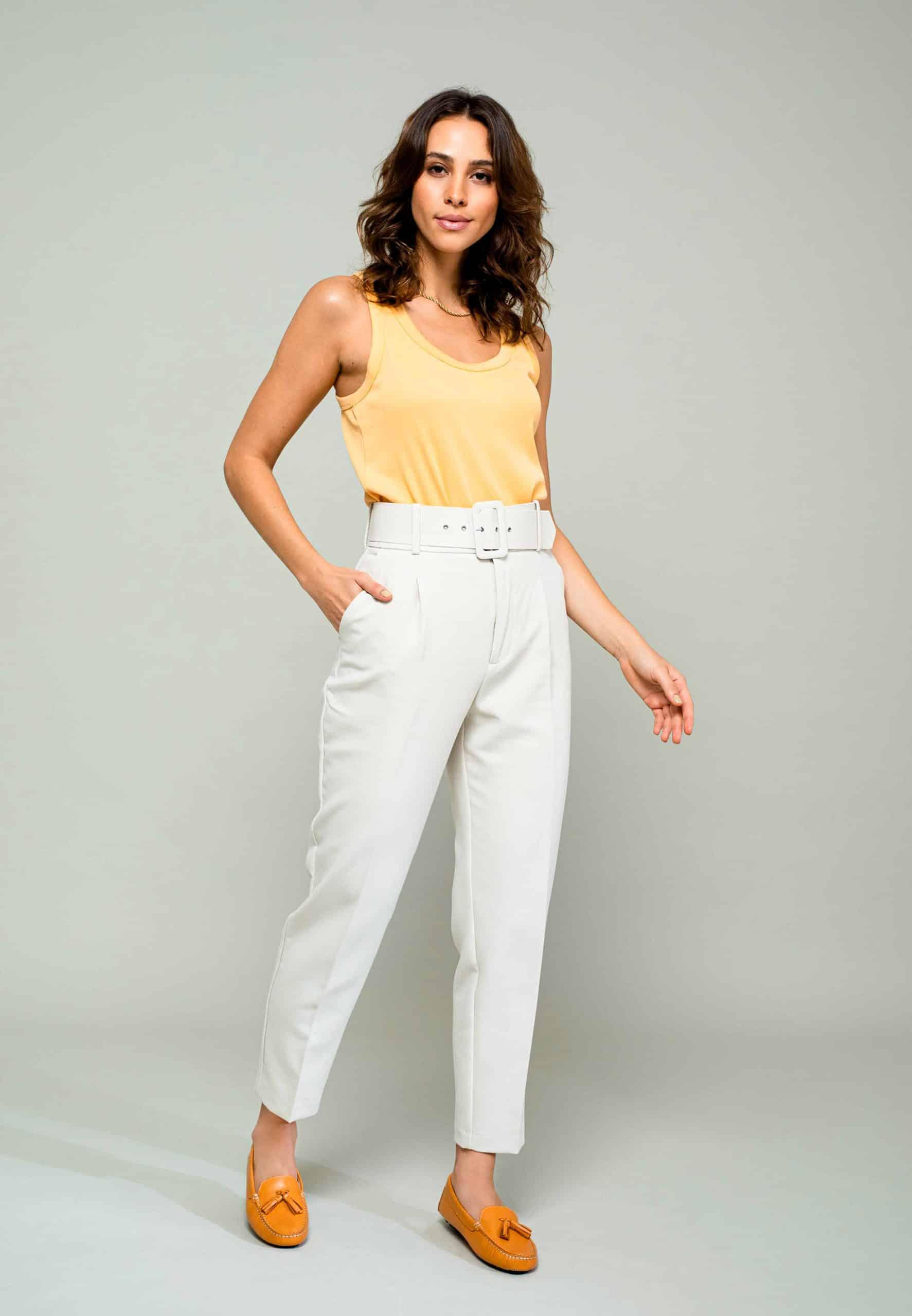 Woman wearing a yellow sleeveless top and white pants on a gray background