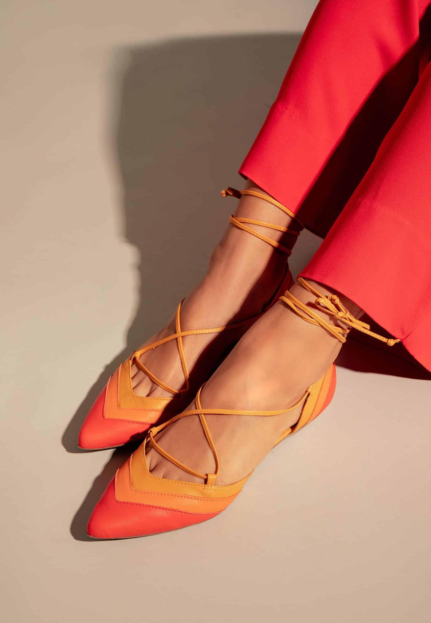 Orange and red flats with criss-cross straps worn with red pants on a neutral background