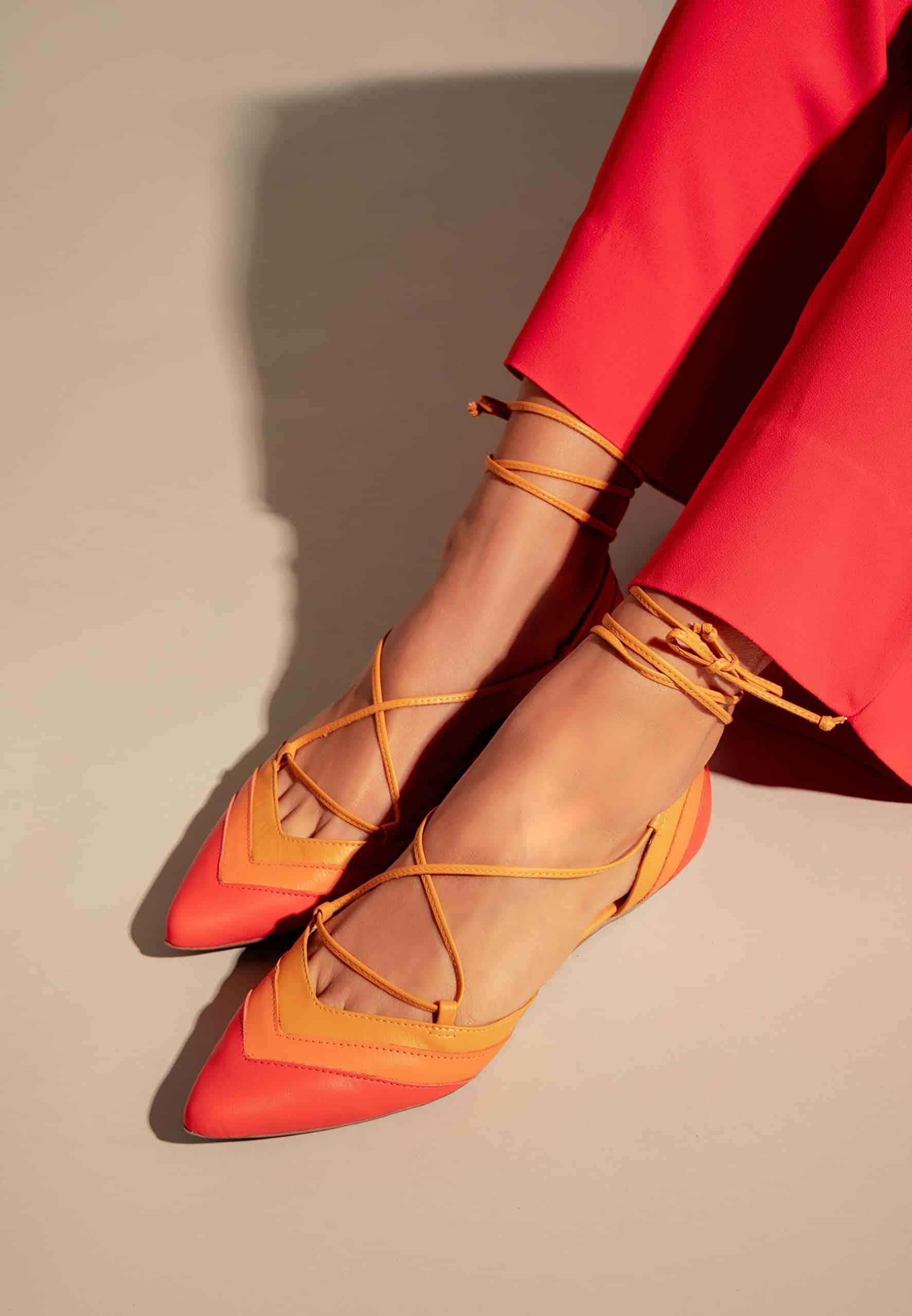 Orange and red flats with criss-cross straps worn with red pants on a neutral background