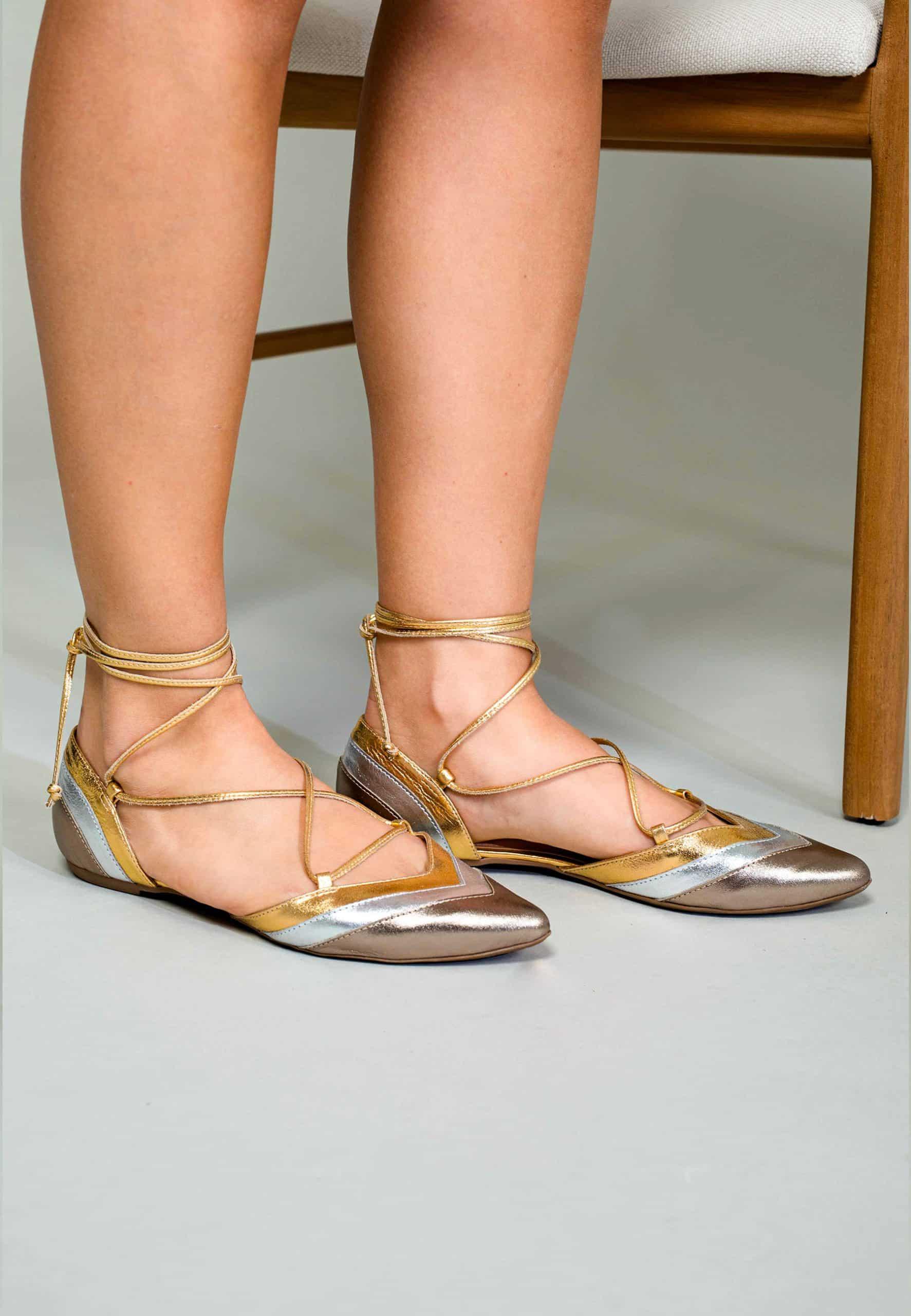 Gold and silver strappy flats worn by a person sitting on a chair.