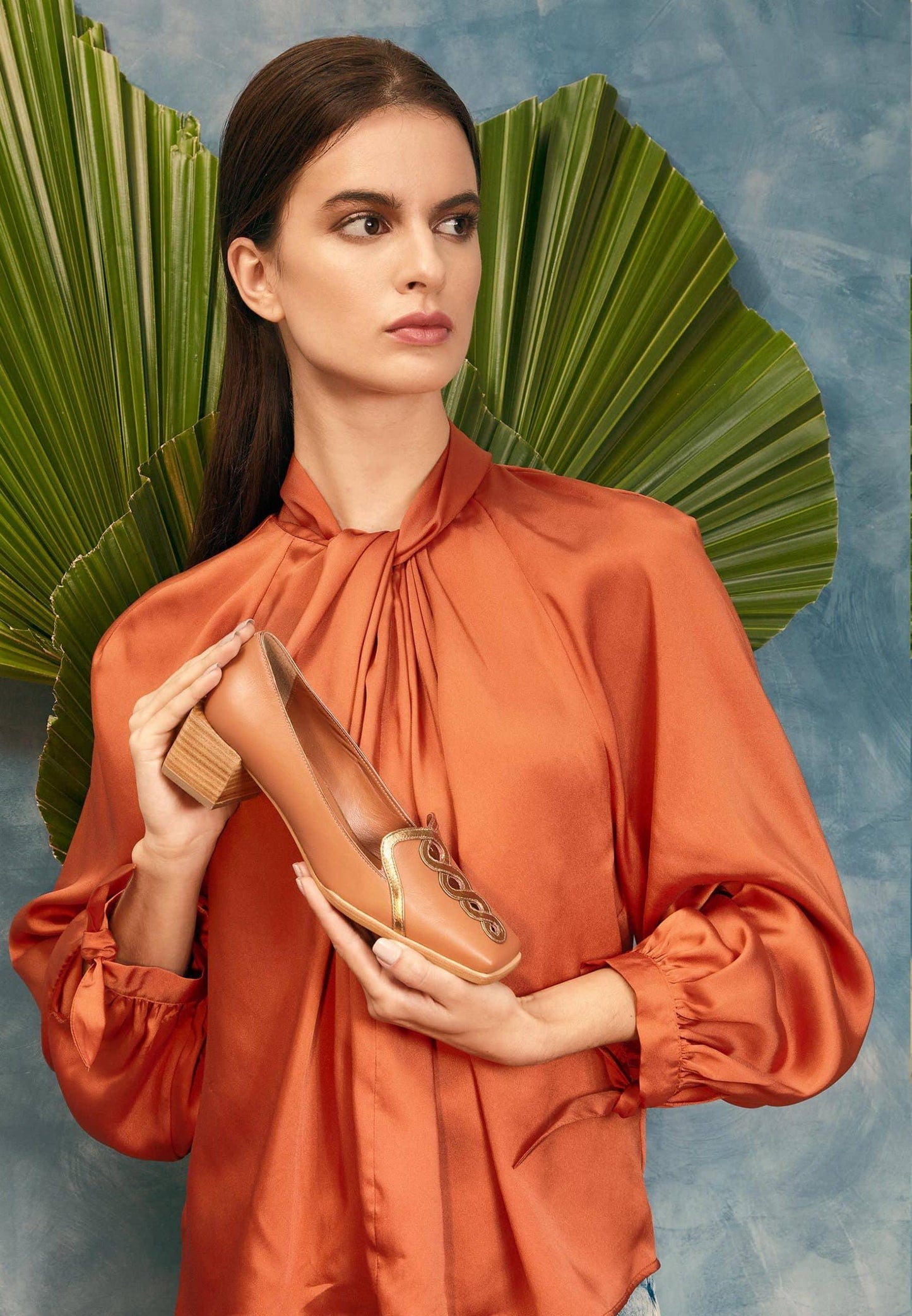 Woman in an orange blouse holding a brown shoe against a decorative background with green leaves.