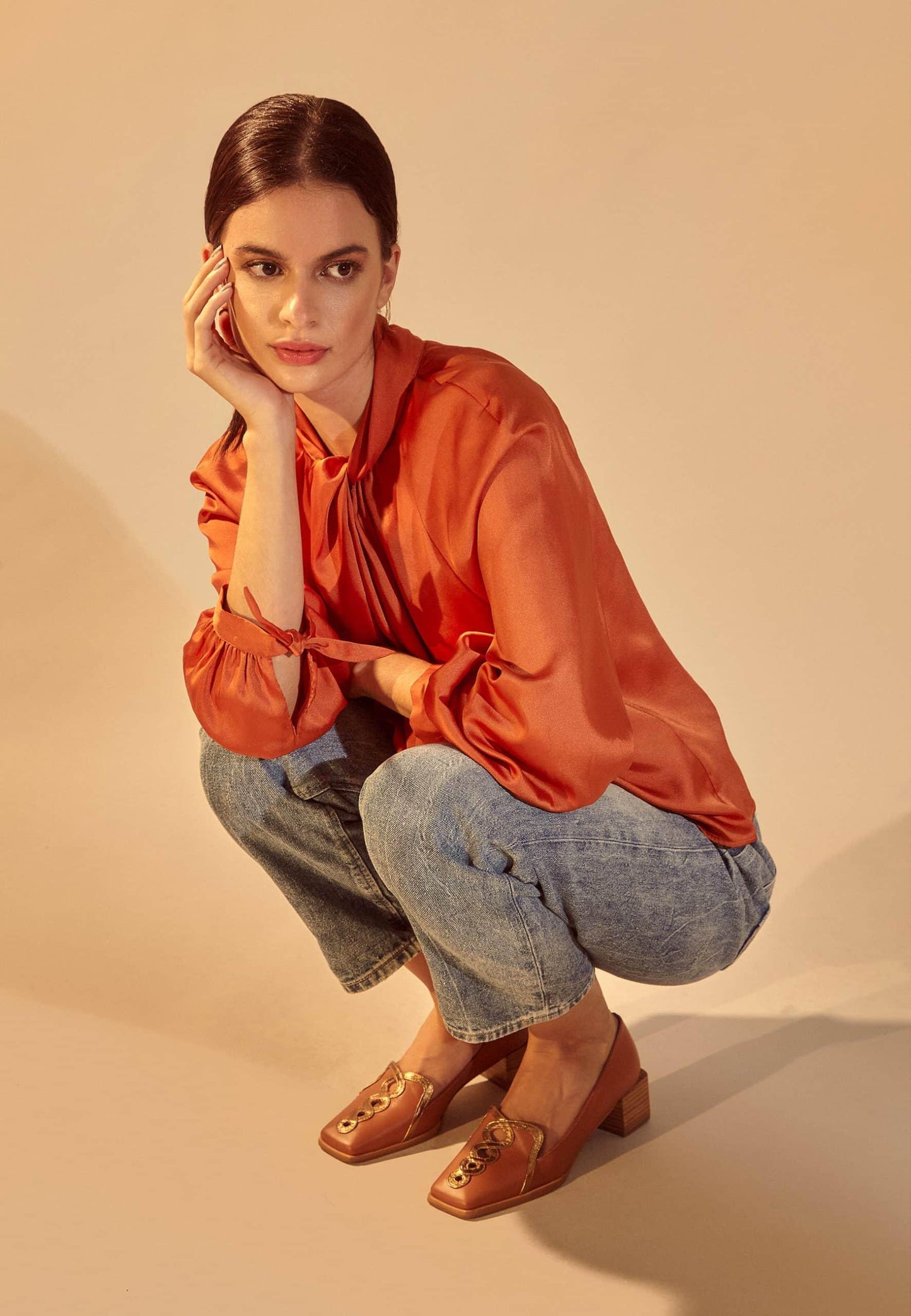 Woman wearing an orange blouse and blue jeans, posing on a beige background