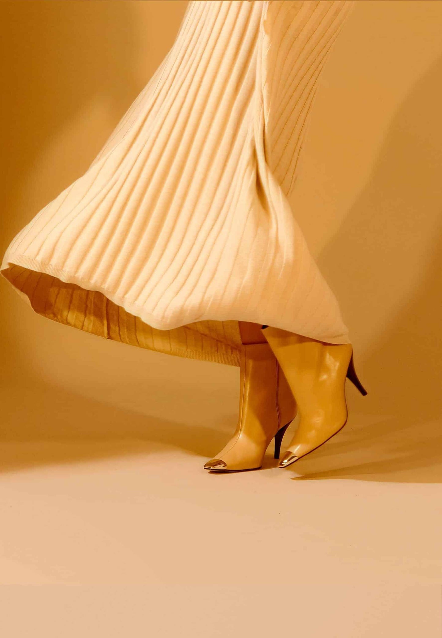 Person wearing mustard yellow boots and a pleated beige skirt against a beige background
