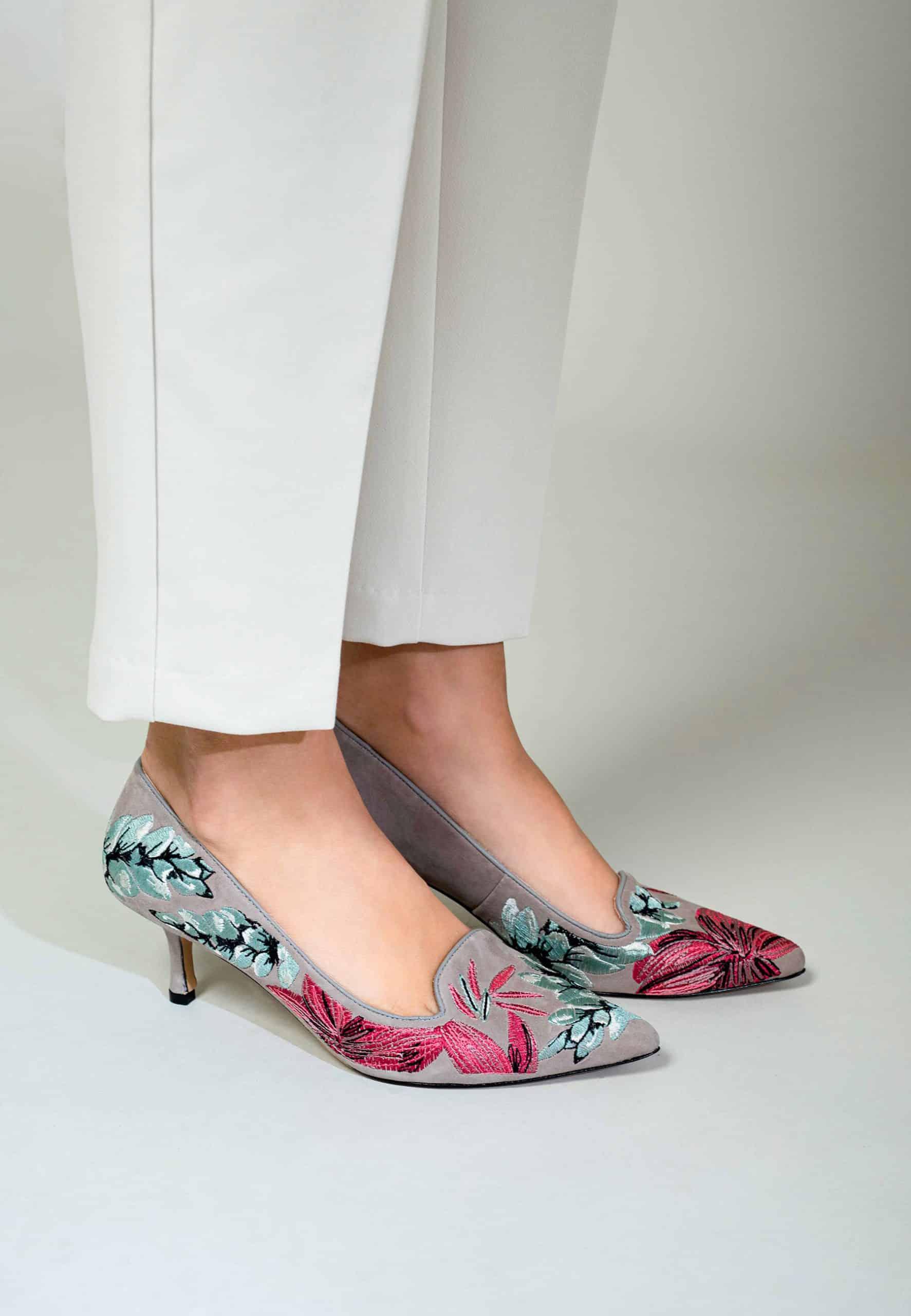 Gray high-heeled shoes with floral embroidery worn with white pants on a light gray background