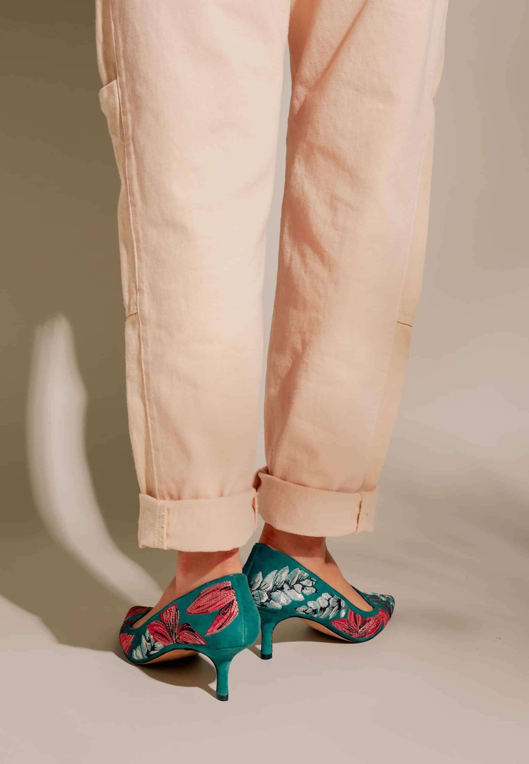 Teal high-heeled shoes with floral embroidery worn with beige pants on a neutral background