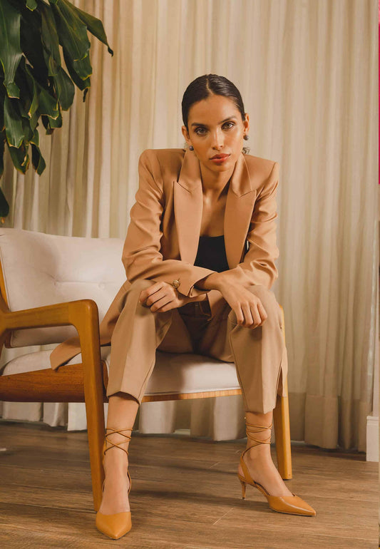 Woman in a beige suit sitting on a chair indoors with curtains and a plant in the background.