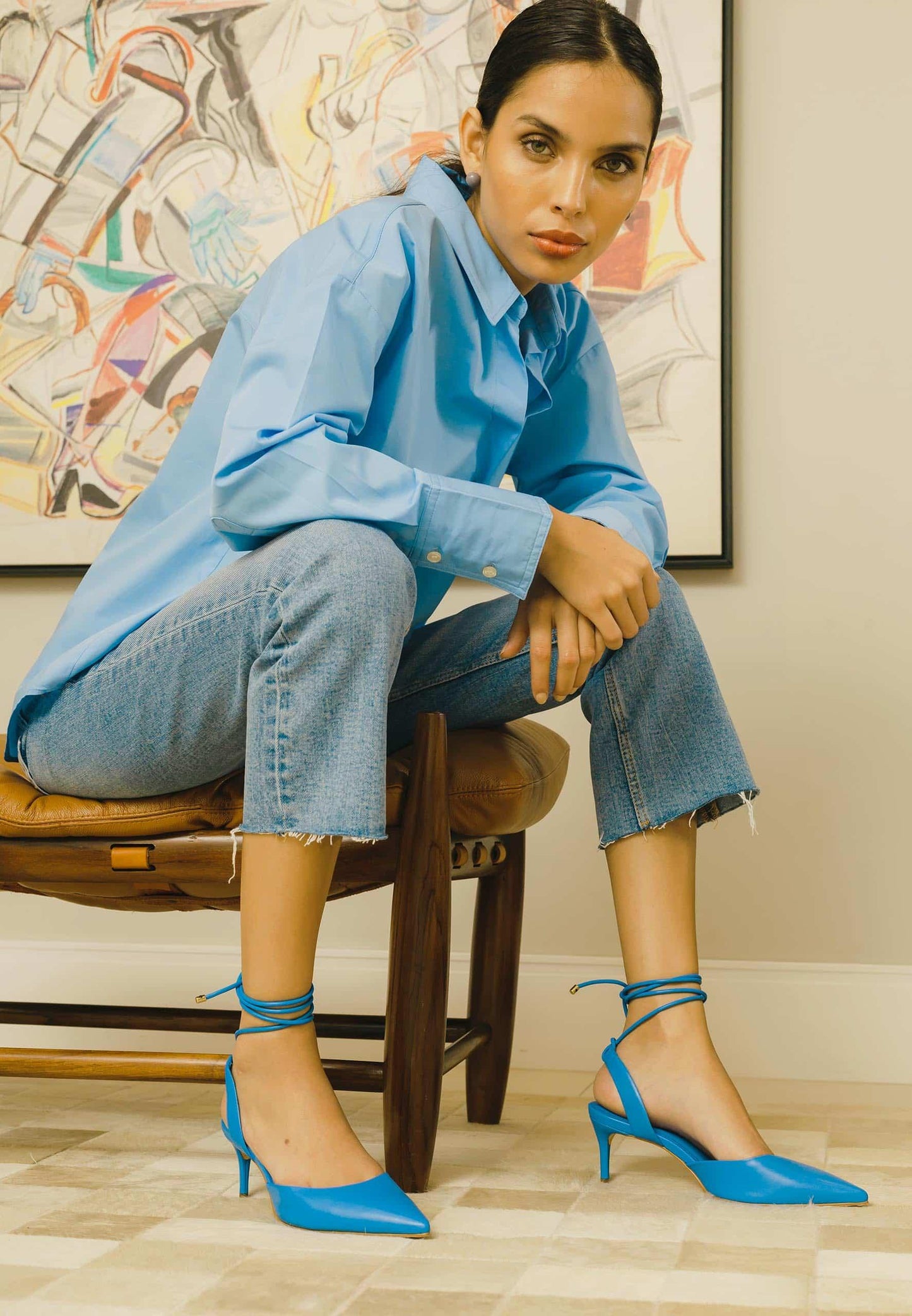 Woman wearing a blue outfit with matching shoes sitting on a chair in a room with abstract art on the wall.